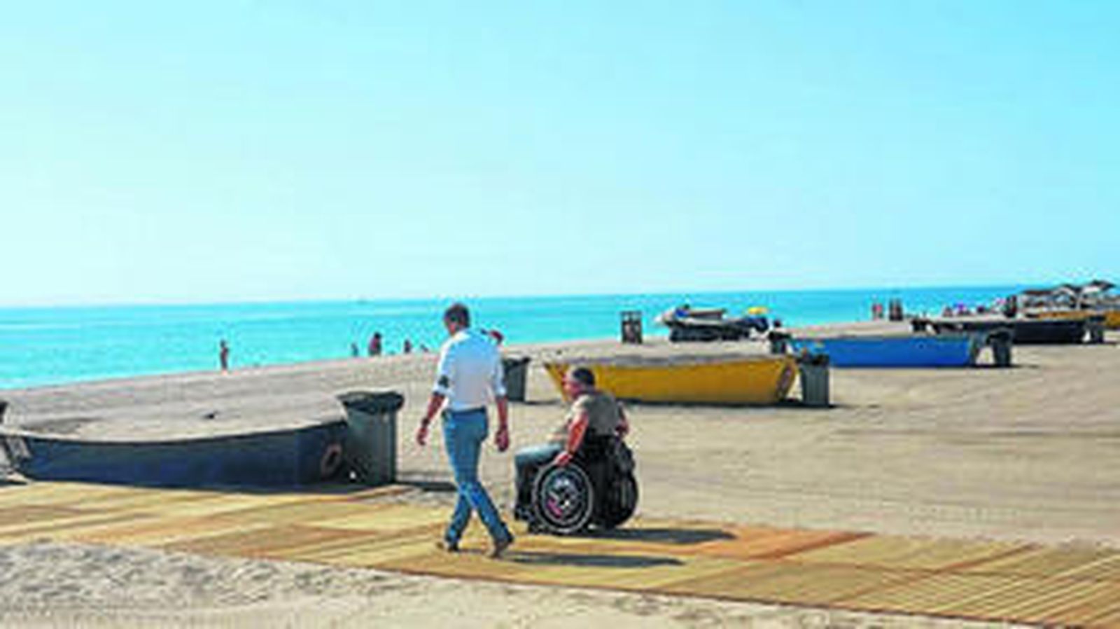 Un usuarios de las playas con discapacidad es acompañado hacia la orilla por la plataforma de madera.