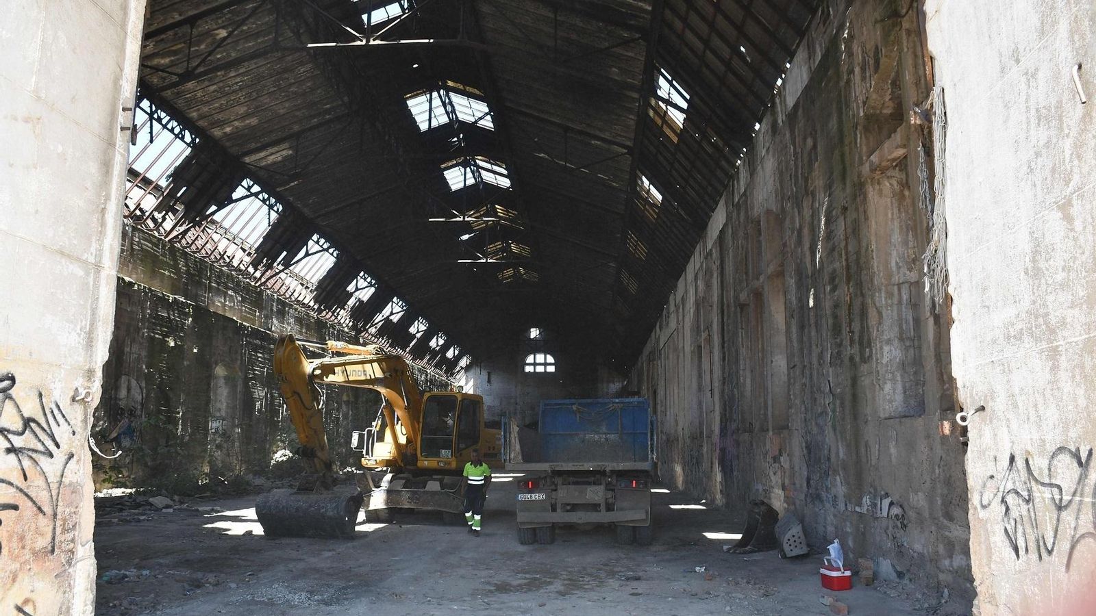 Un operario junto a una excavadora y un camión en el interior del edificio.