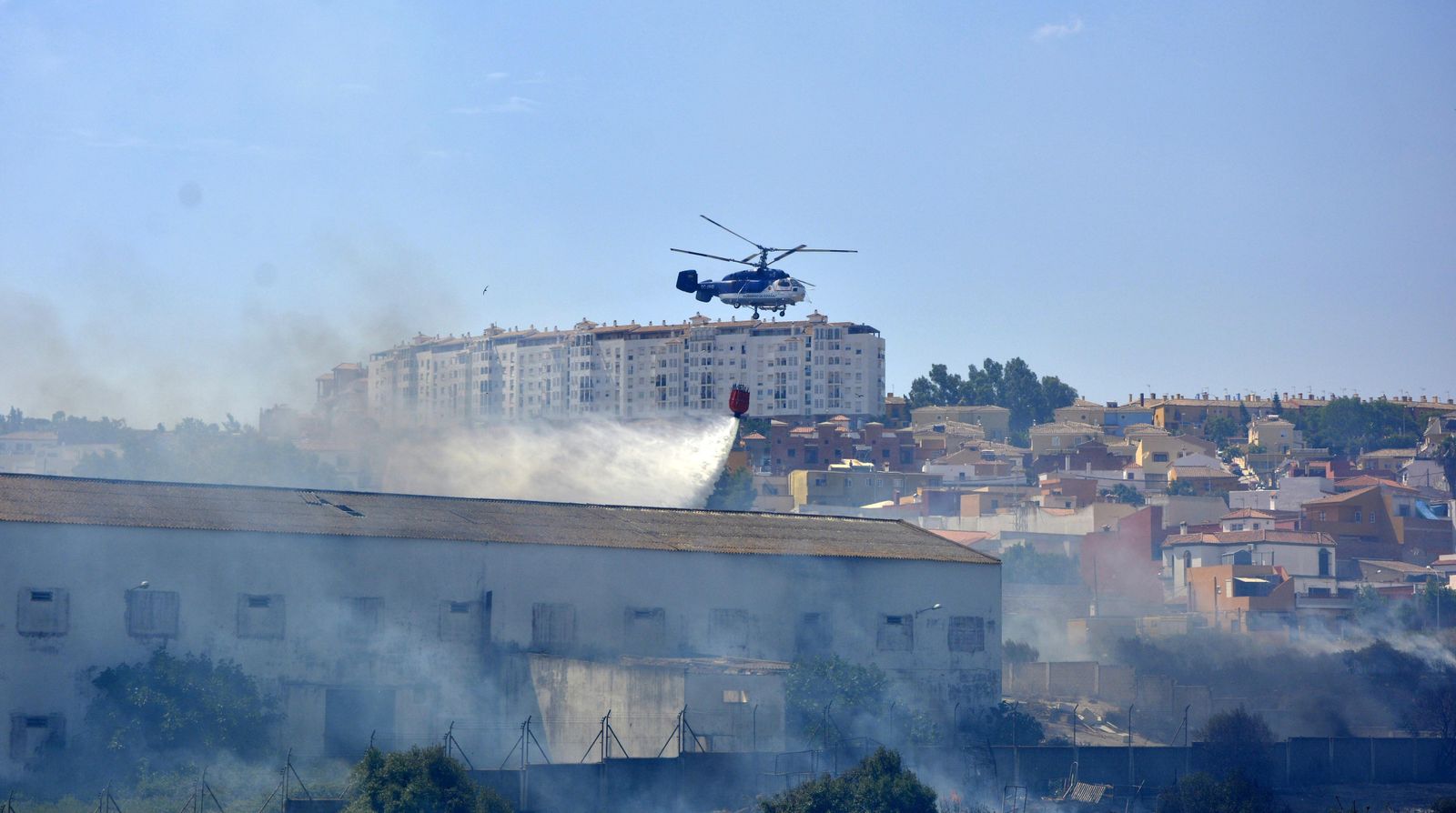 Un helicóptero refresca la zona del incendio, muy cercano a las viviendas en la barriada La Granja, en Algeciras.