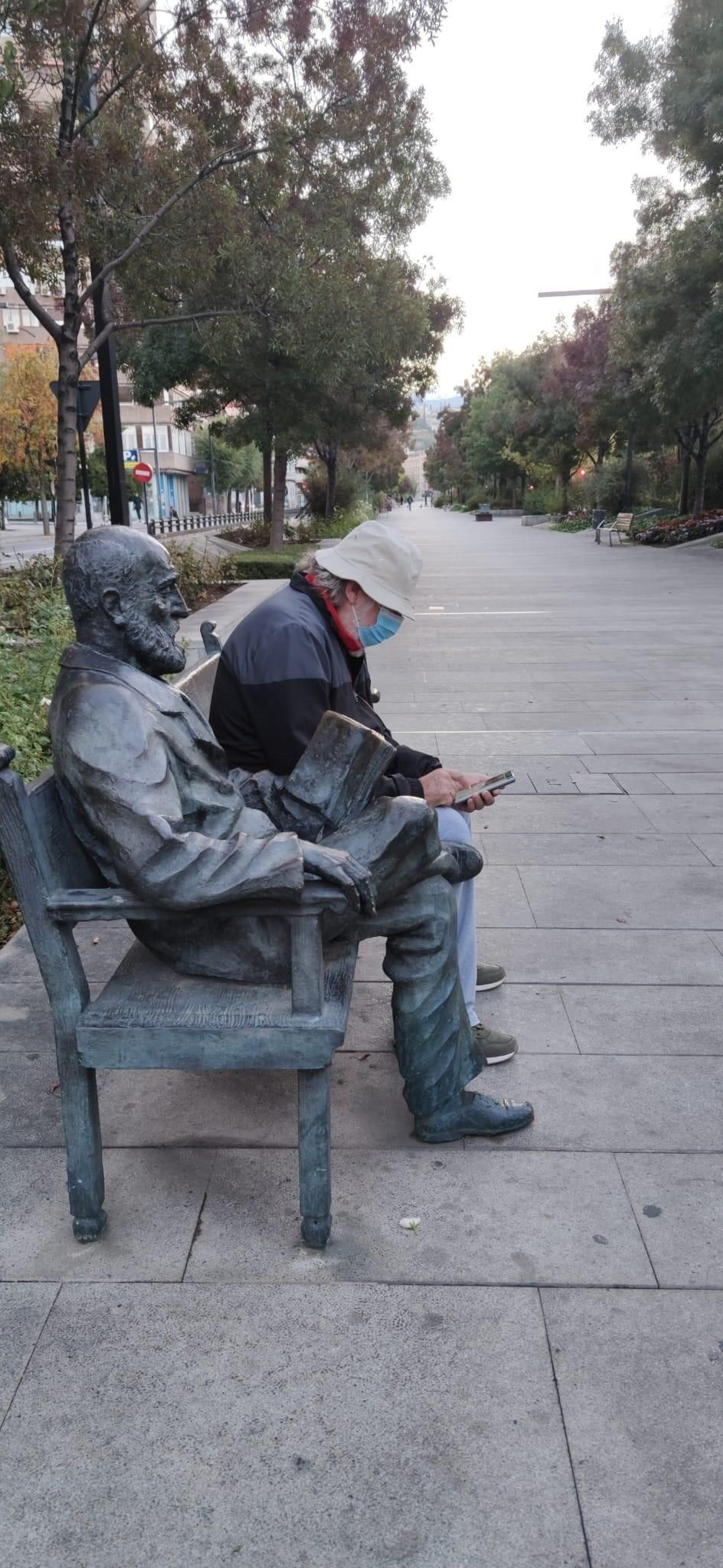 Pedro Antonio de Alarcón lee un libro mientras que su compañero de banco observa el móvil