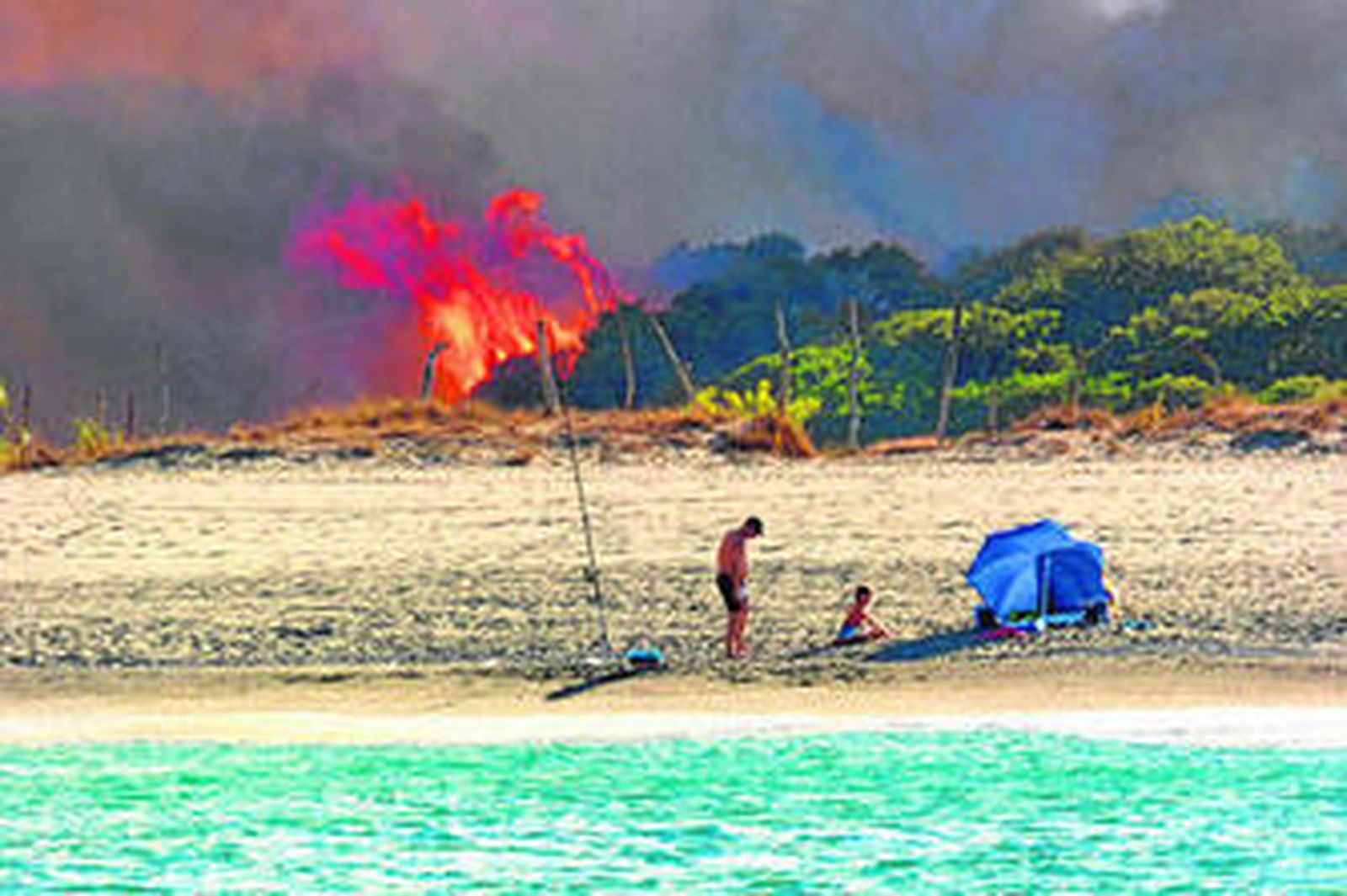 Unos bañistas permanecen ajenos a la columna de llamas que se acerca a la playa.
