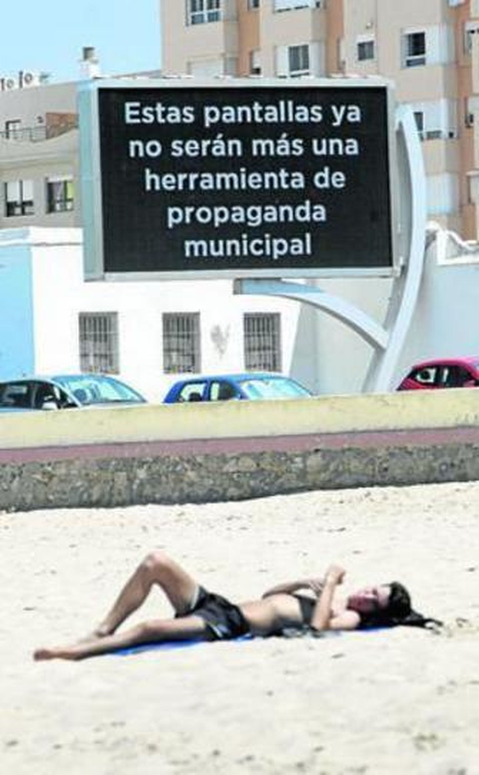 Un hombre toma el sol en la playa con la pantalla LED y su anuncio al fondo.