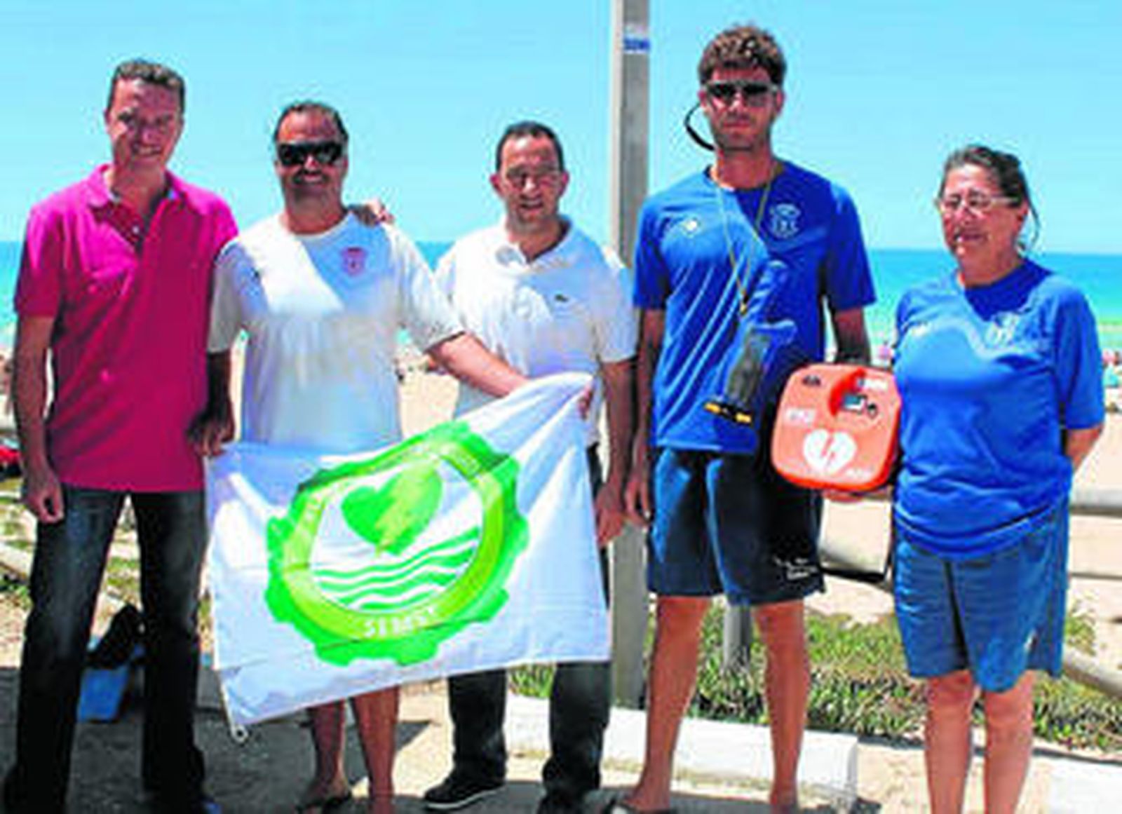 Miembros del equipo sanitario de la playa muestran la bandera del certificado y el desfibrilador.