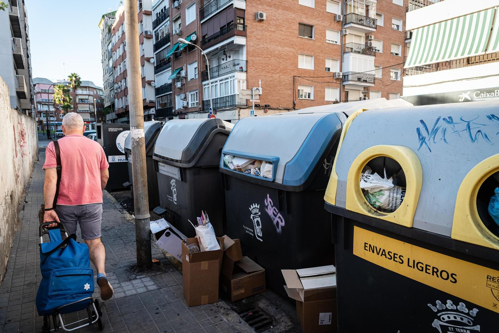 Cajas de cartón en una acera llena de manchas junto a un contenedor de papel repleto.