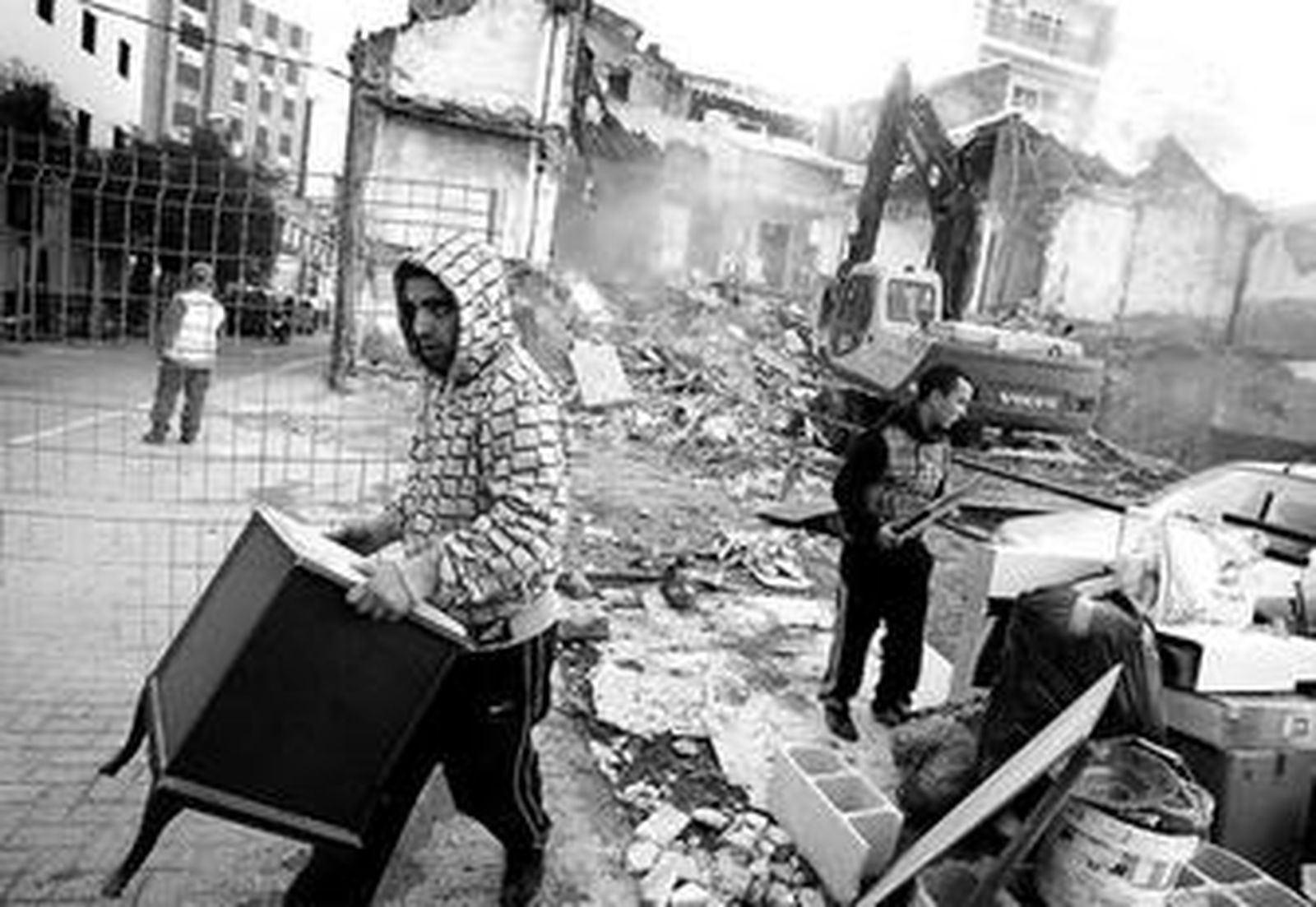 Algunos vecinos desalojados portan sus enseres mientras la máquina destroza el edificio.