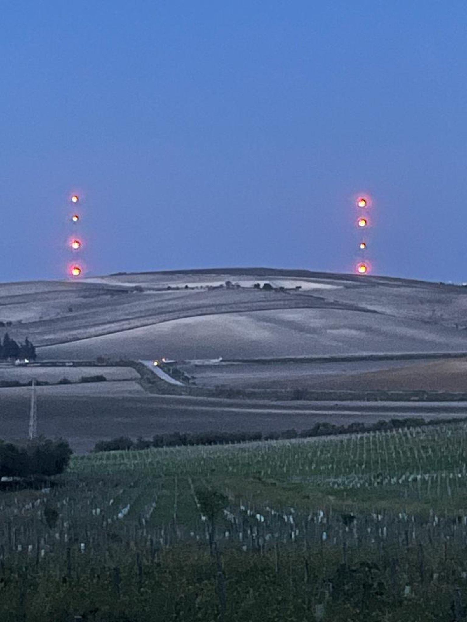 Cultivos y viviendas en la zona del viñedo afectada por el parque eólico con las balizas iluminadas al fondo.