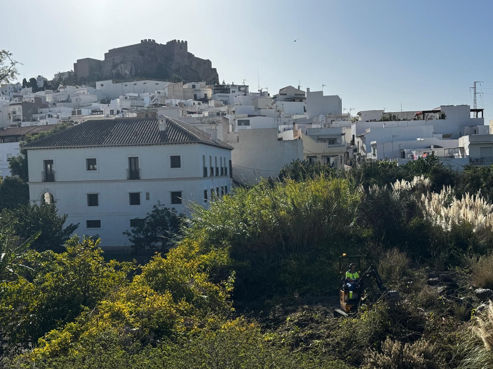 Un operador desbroza el solar donde irá ubicado el depósito, con el Castillo y el depósito de Antequera al fondo
