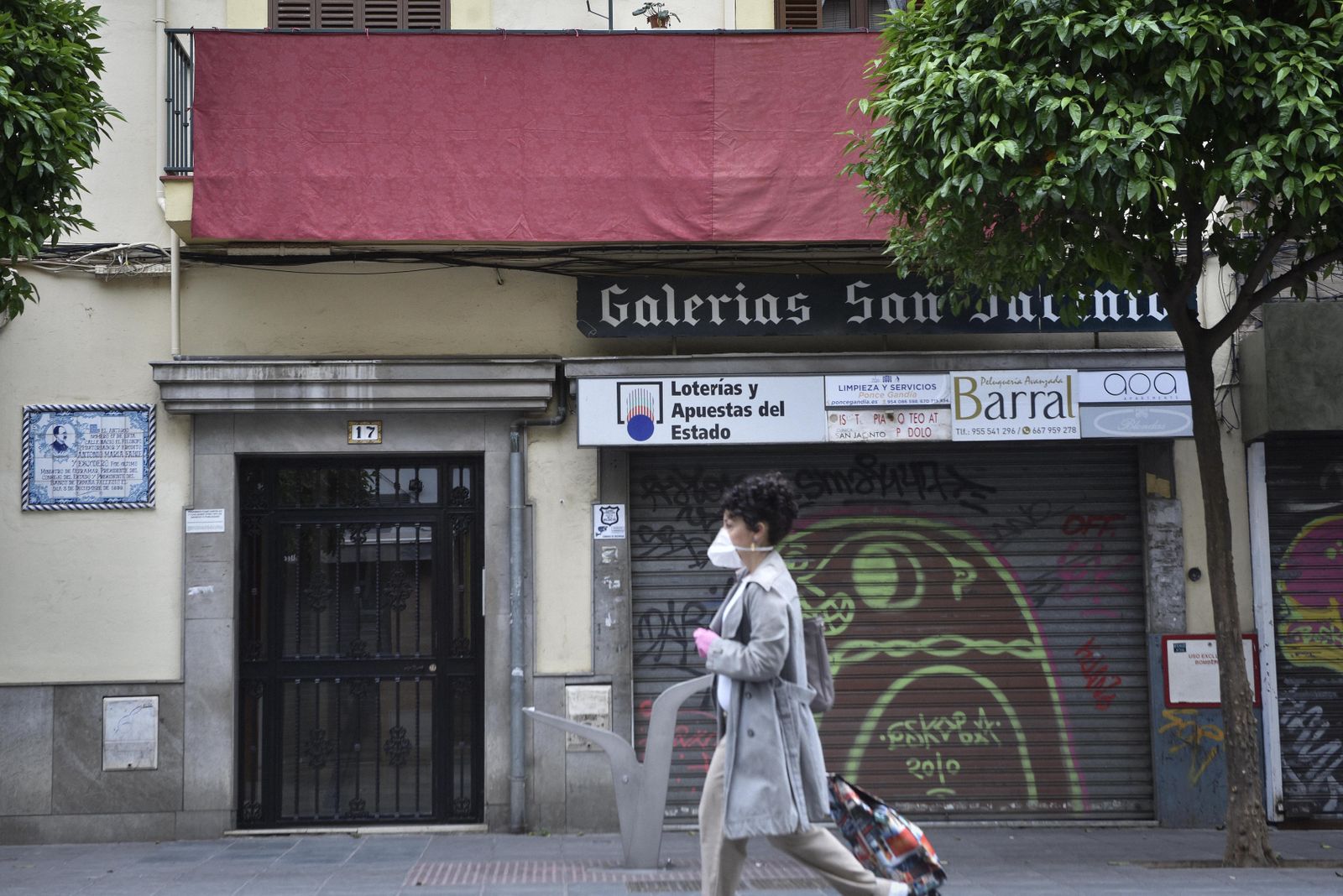 Una mujer con mascarilla y el carro de la compra pasa junto a un balcón engalanado.
