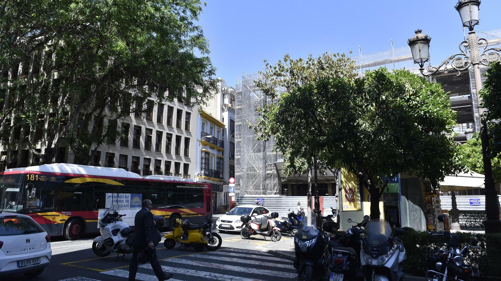 Un peatón cruza la plaza, con un autobús al lado y entre motos aparcadas y con las obras del hotel, al fondo.