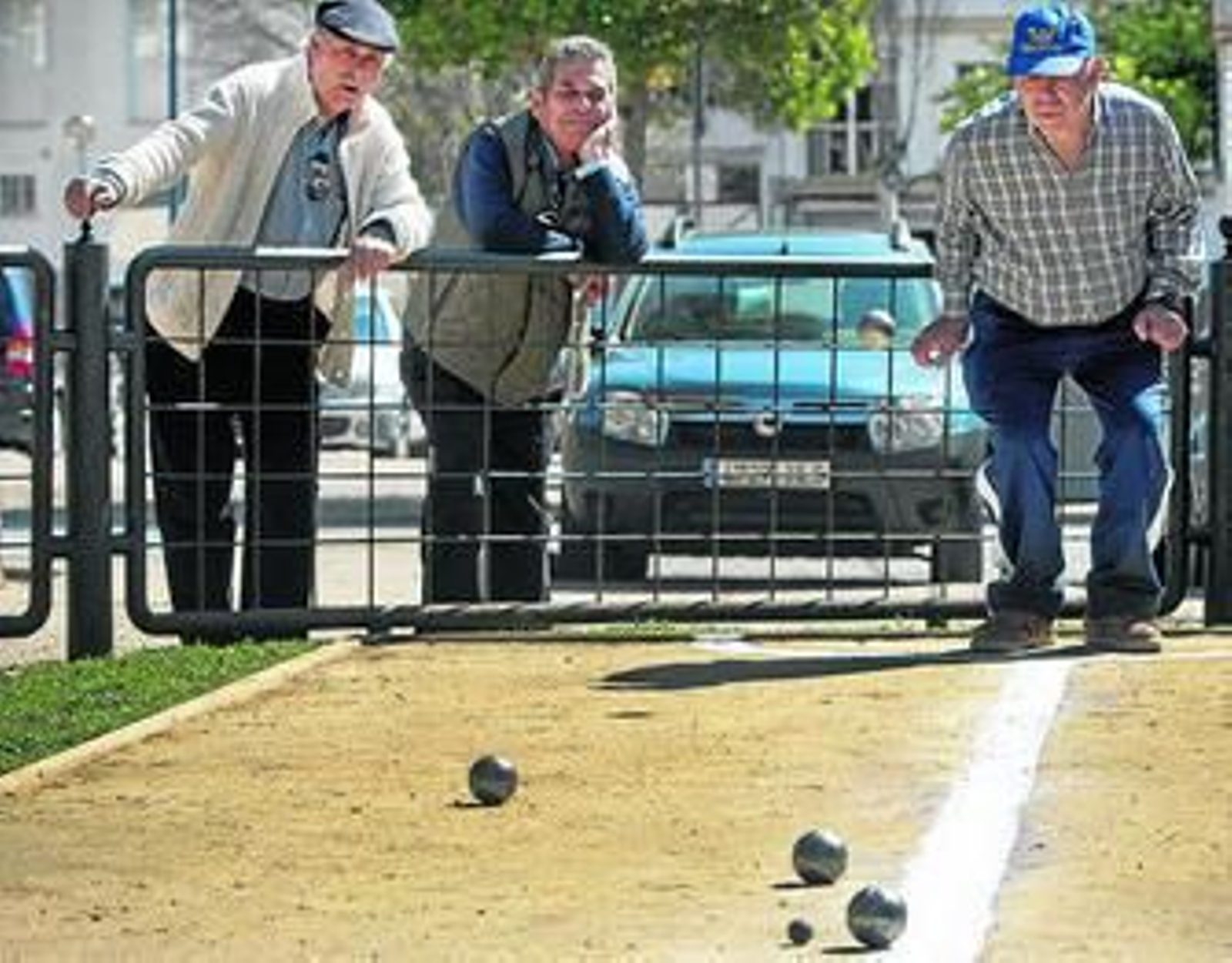 Dos isleños observan cómo un jugador lanza la bola en el juego de petanca.