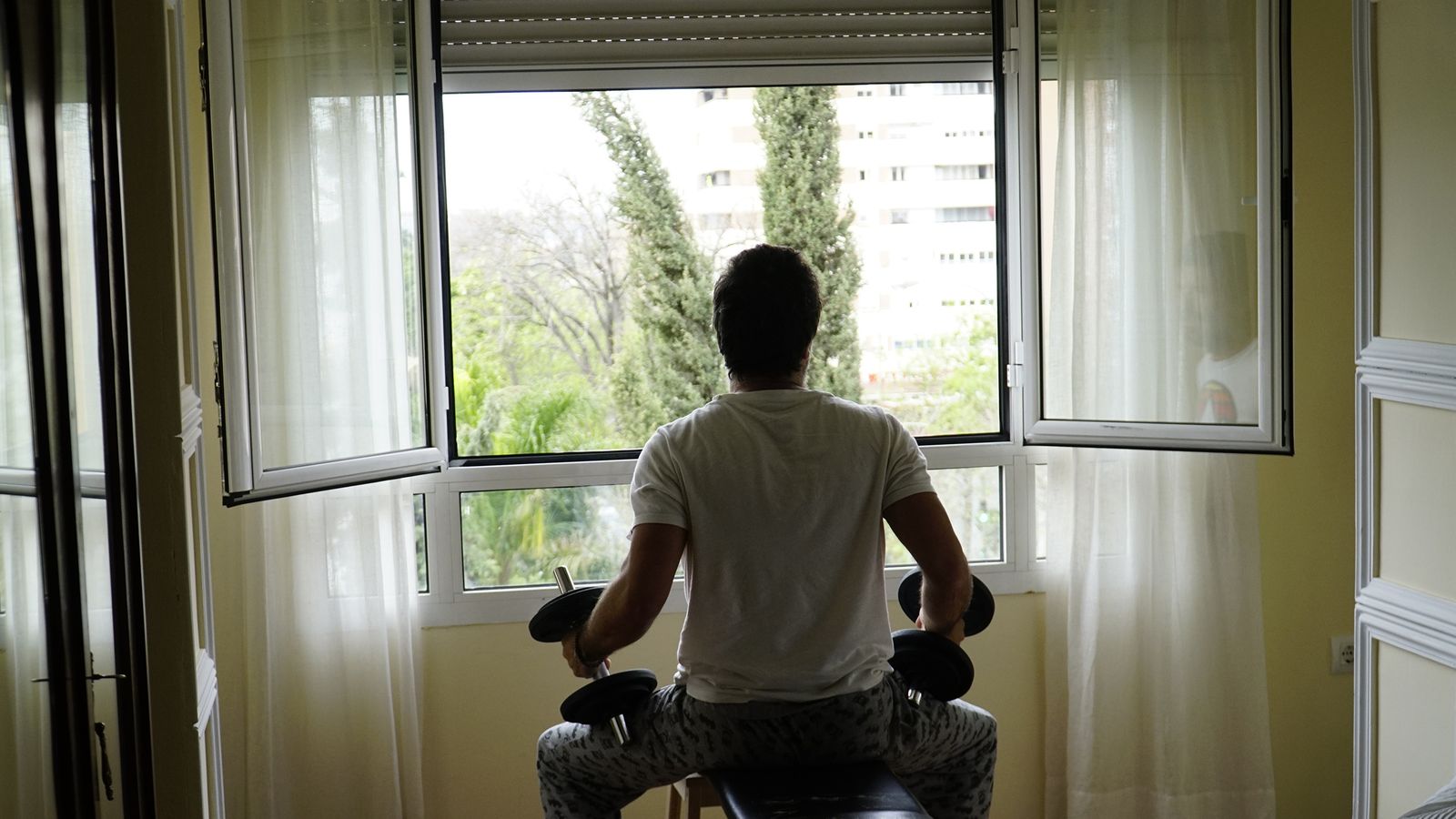 Un hombre, pesas en mano, observa el exterior desde su ventana.
