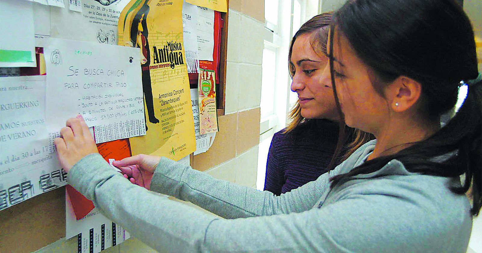 Alumnas ante un tablón con anuncios de alquileres de pisos en una facultad universitaria.