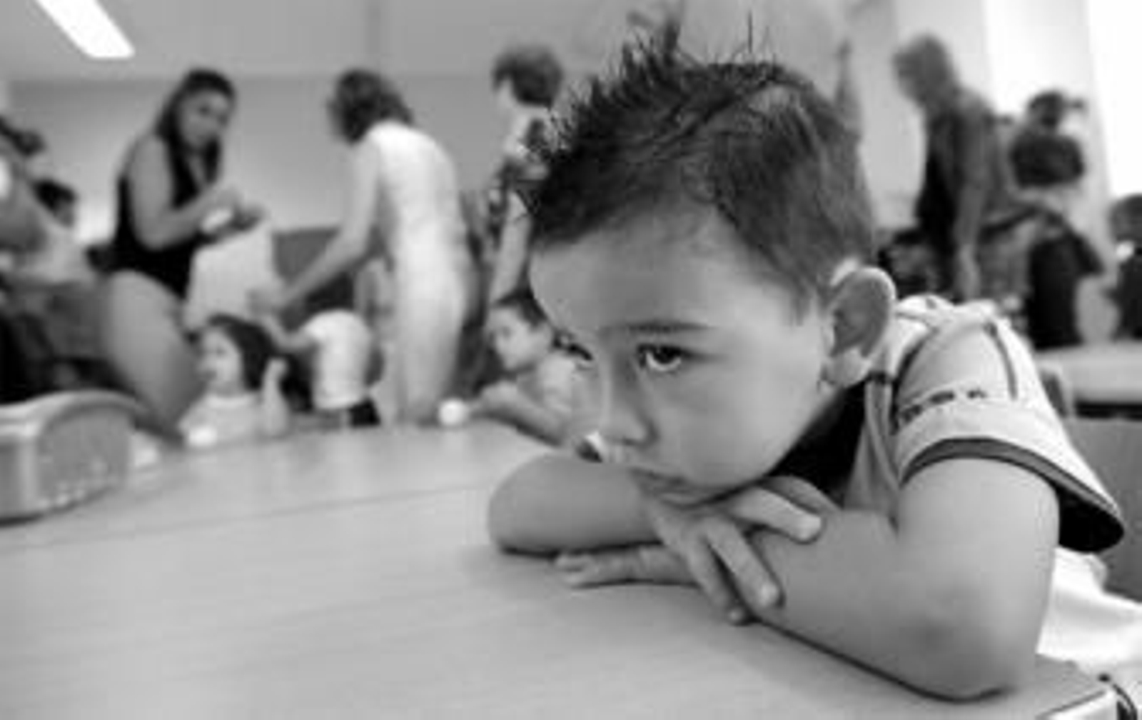 Un niño reflexiona en su pupitre, mientras al fondo un grupo de madres trata de poner orden para realizar una actividad.