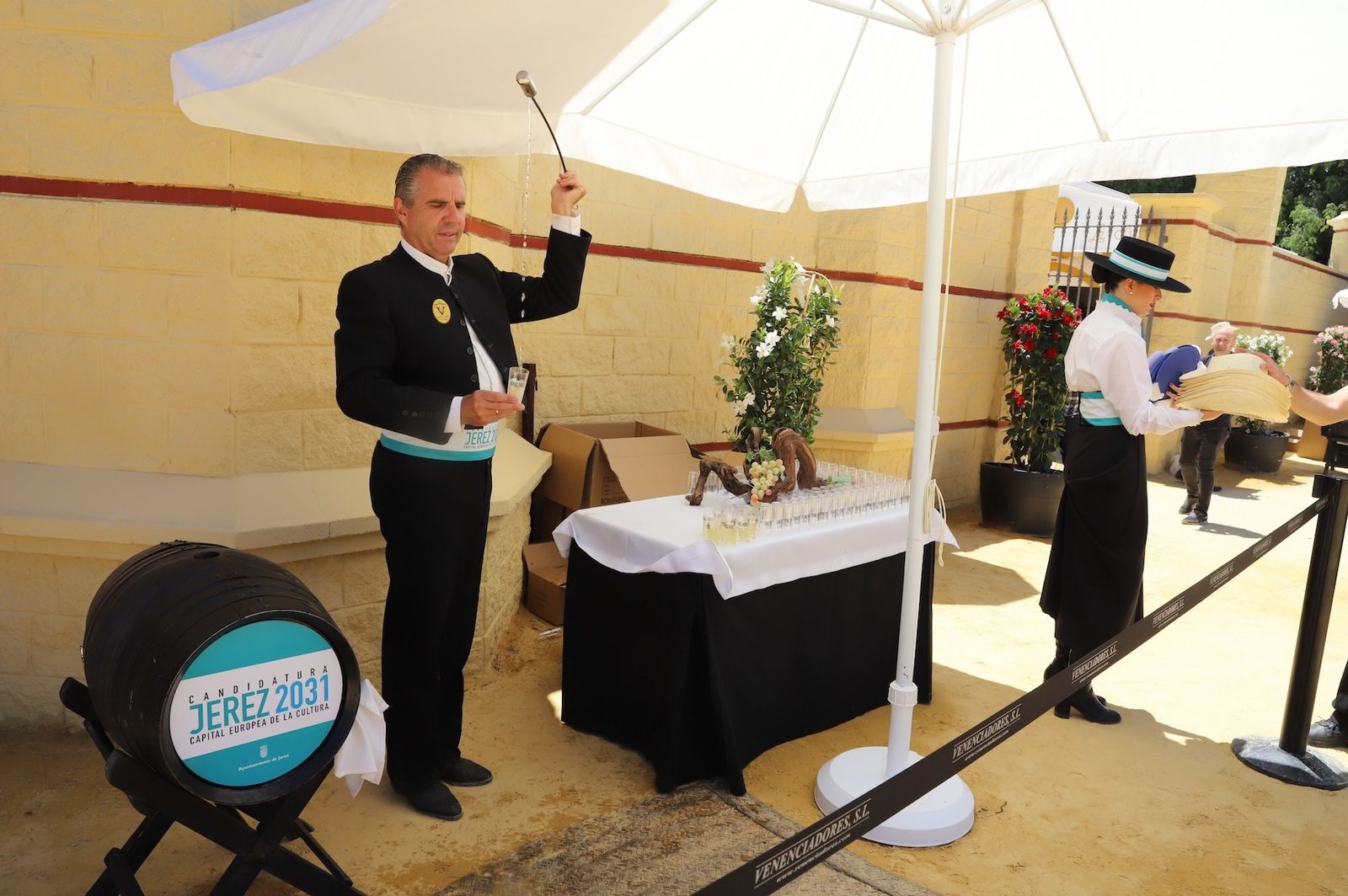 Un venenciador repartiendo vino y una azafata entregando merchandising alusivo a la capitalidad cultursa desde el Punto de Información Turística, ubicado en la fachada principal de la Feria de Jerez