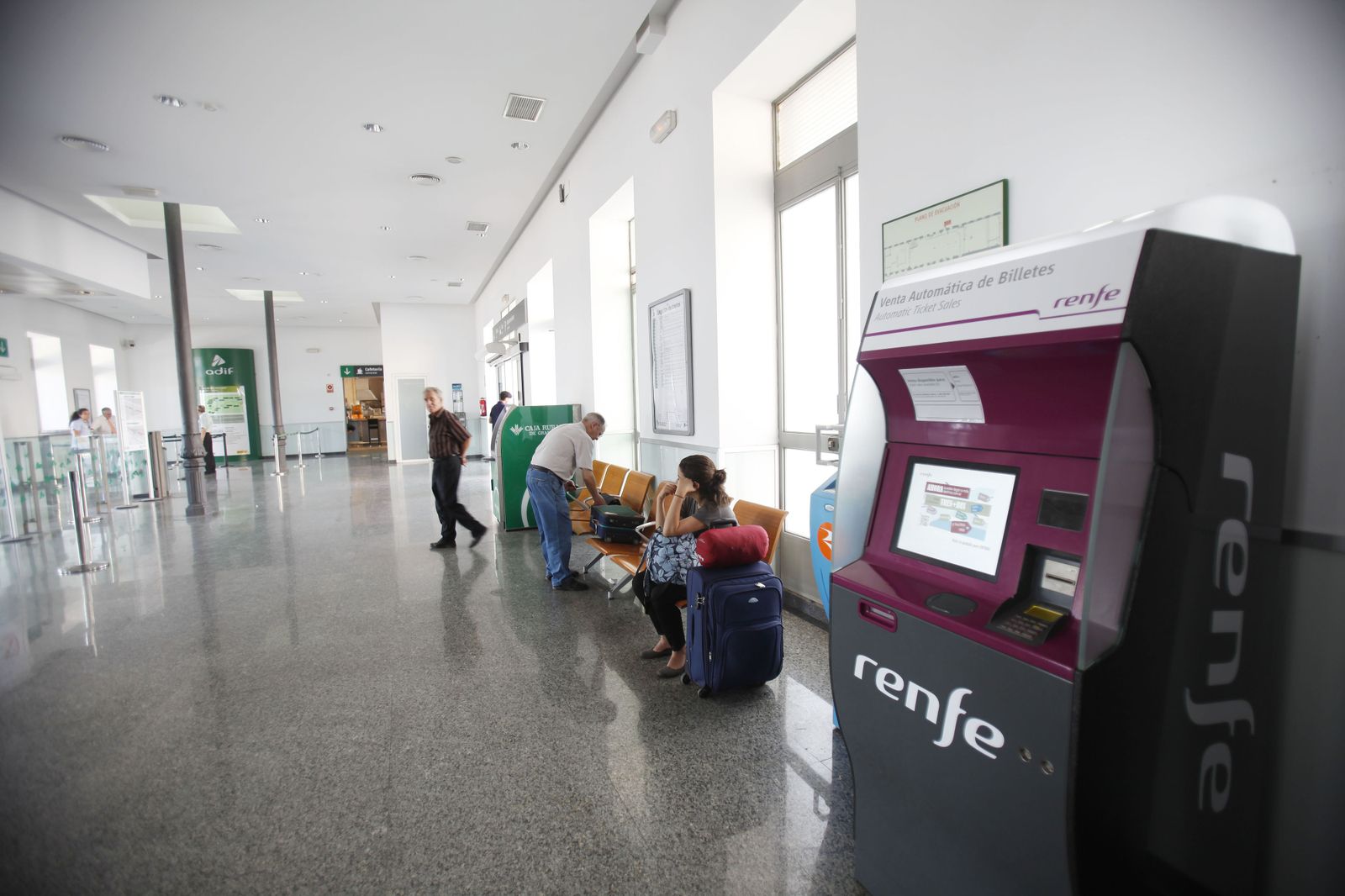 Pasajeros esperan en una estación de Adif junto a un dispensador de billetes de Renfe.