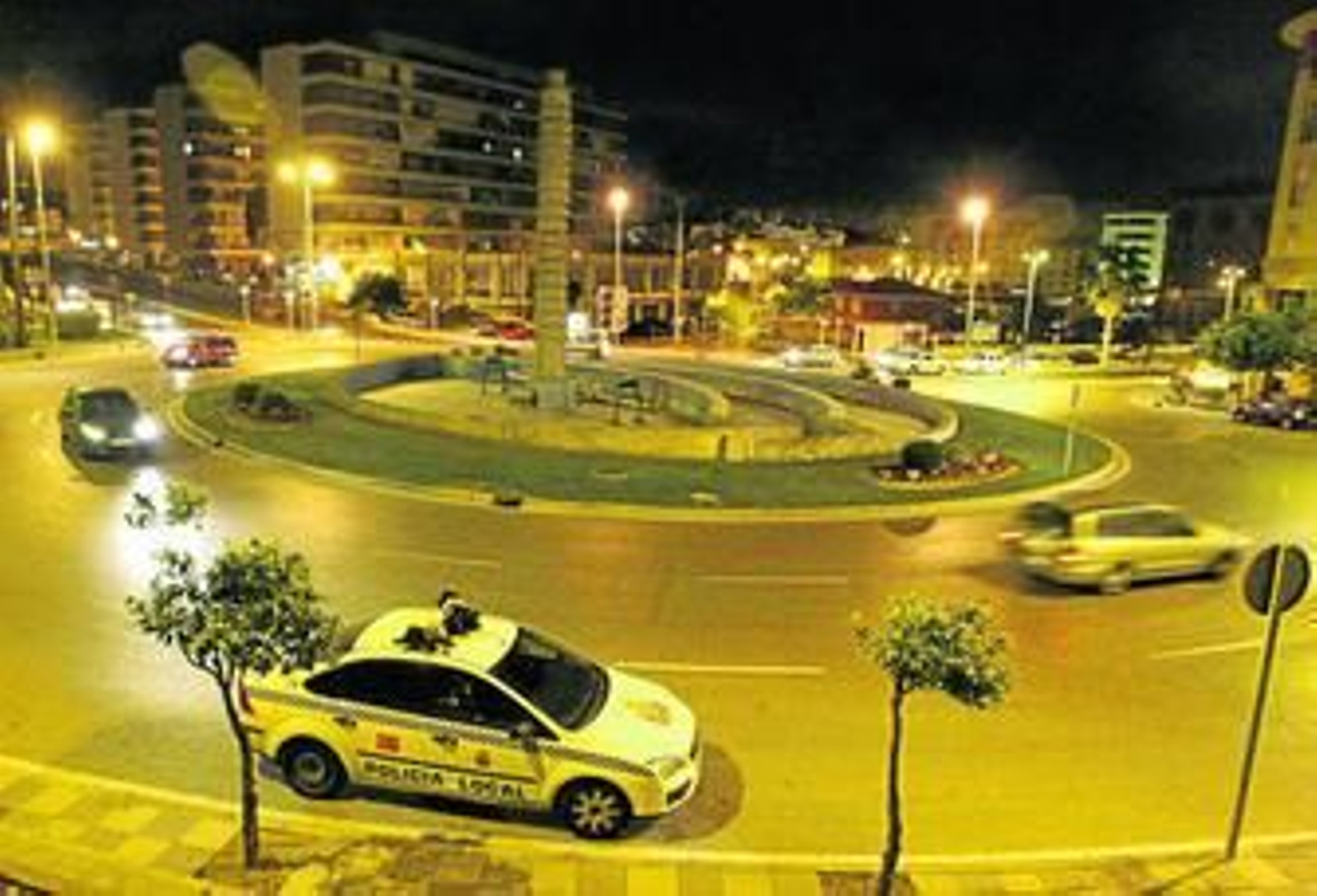 Imagen nocturna de la rotonda con el monumento al milenio, en Algeciras, siendo rodeada por varios coches.