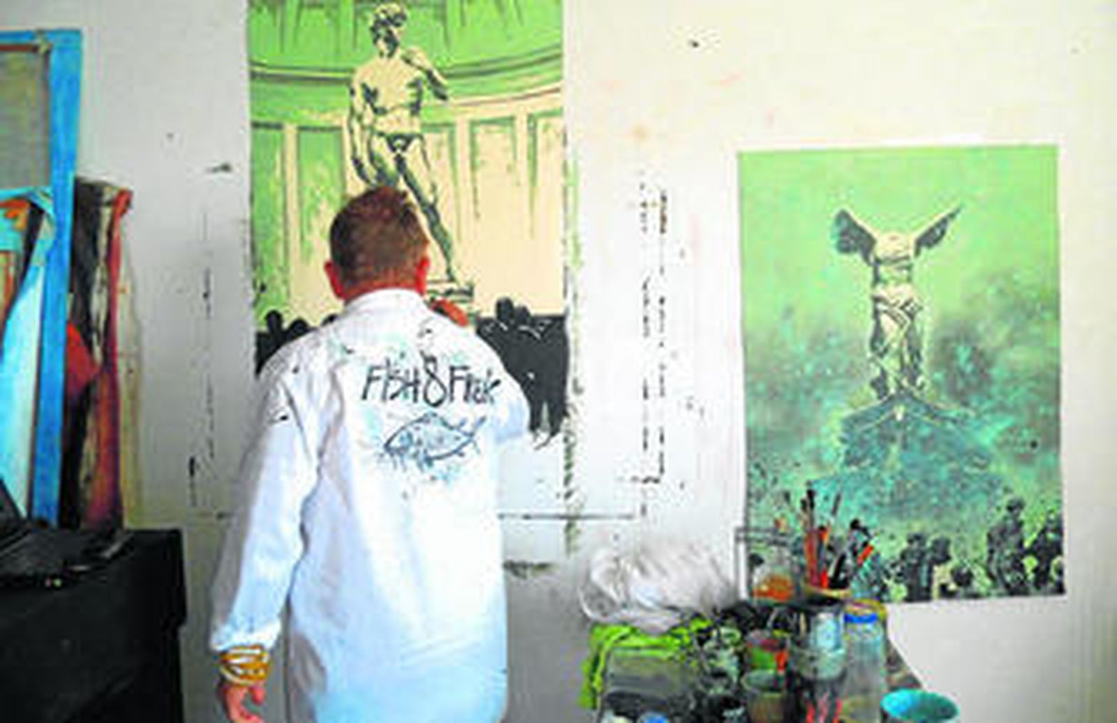 El artista, pintando en su estudio, con una camisa donde se puede leer el título de la exposición.