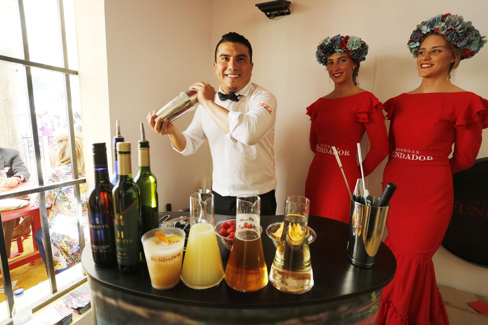 Ricardo González, bartender de FIZZ, elabora uno de los cócteles de Harveys junto a dos azafatas de Fundador en la caseta de la bodega en la Feria.