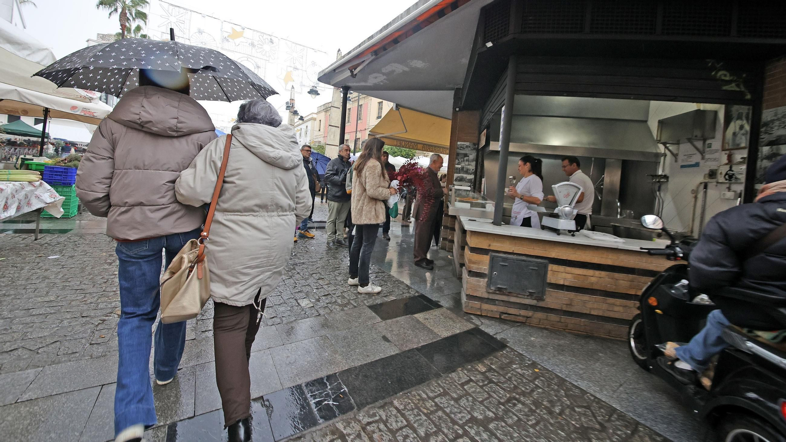 Dos personas caminan con paraguas cerca del puesto de churros de la plaza de Abastos de Jerez mientras llueve.