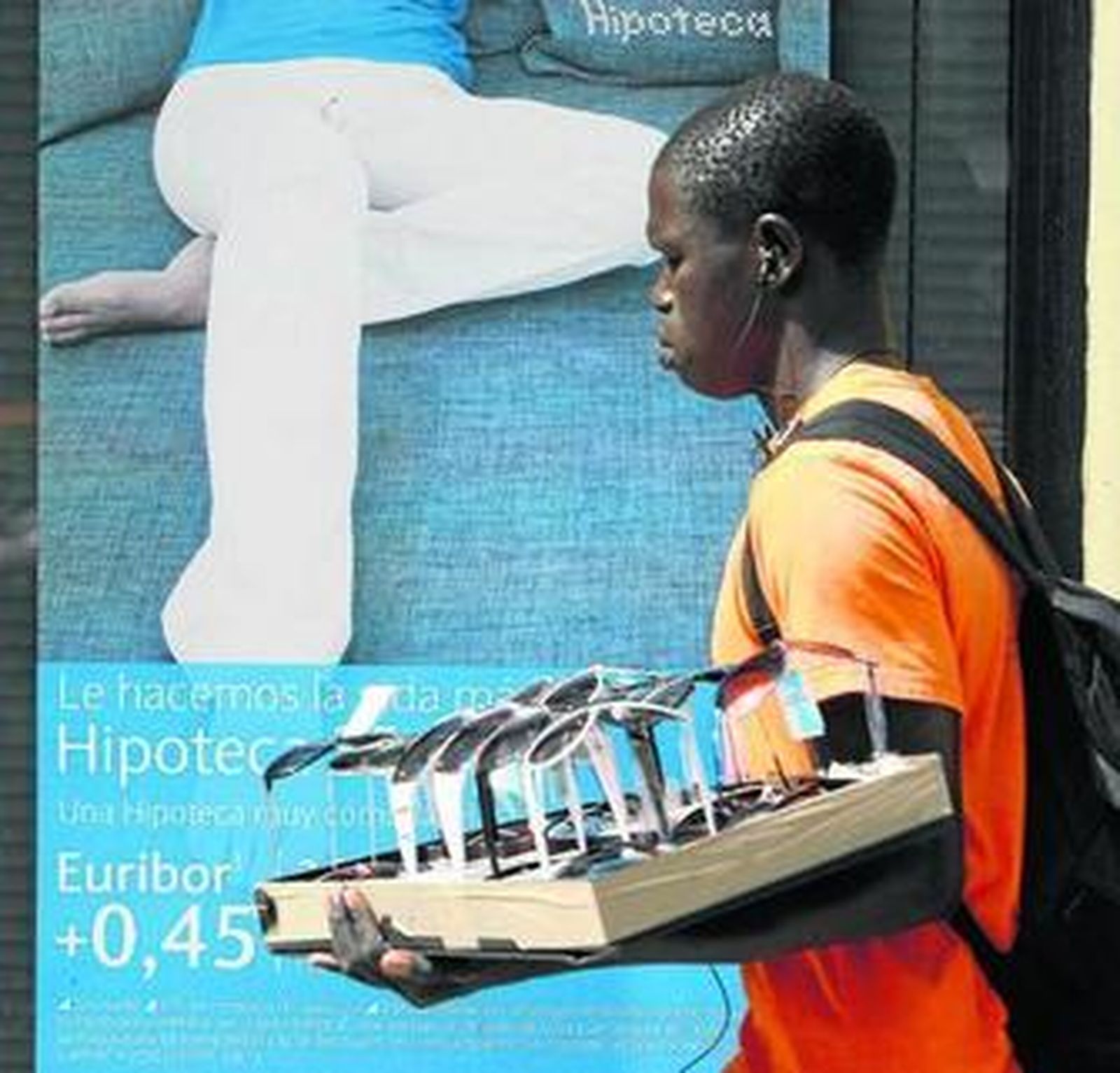 Un vendedor ambulante de gafas de sol pasa por delante de una sucursal bancaria que oferta hipotecas, en el centro de Cádiz.