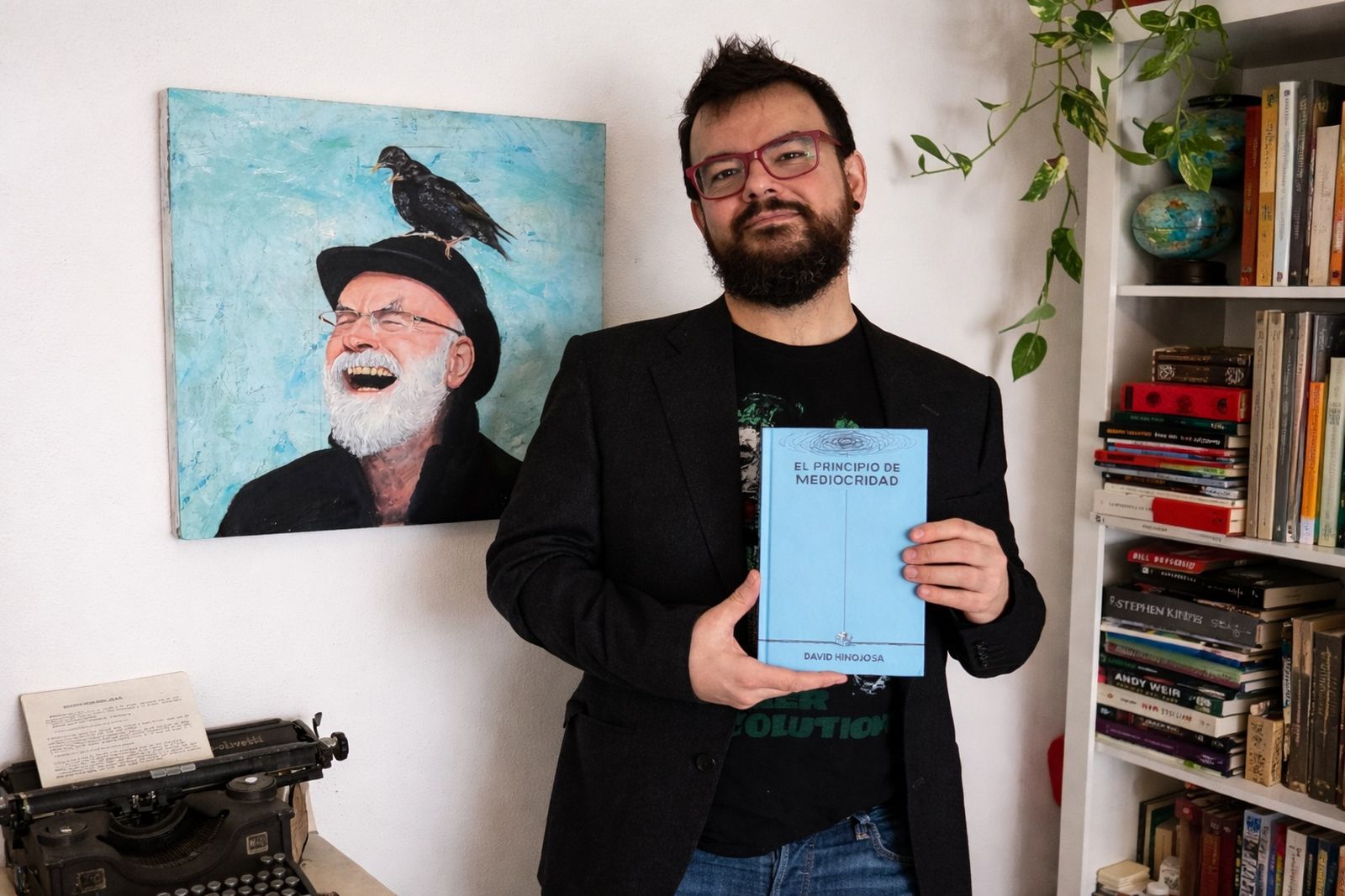 David Hinojosa con un ejemplar de su nueva novela junto a un óleo con el retrato de Terry Pratchett, uno de sus autores fetiche, hecho por él mismo.