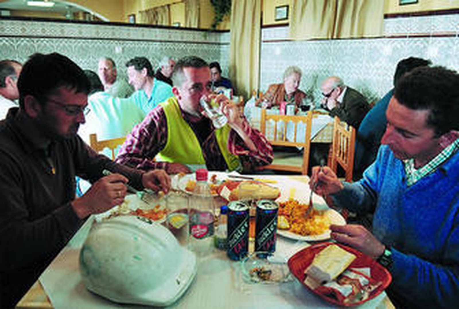 Tres trabajadores de la obra, comiendo en el restaurante Sancho Panza, con el casco encima de la mesa. Tras ellos, más.