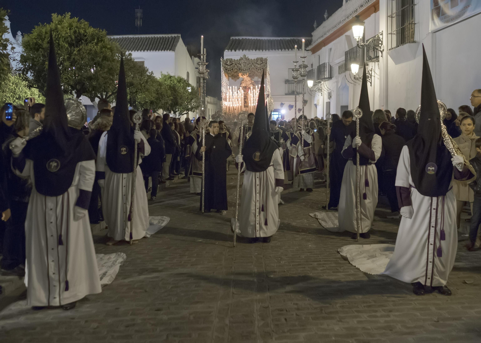 La cofradía de la Soledad de Olivares cruza la Plaza de España de este municipio aljarafeño. Los nazarenos arrastran la cola de sus túnicas. Al fondo, el elegante palio de la Virgen de los Dolores.