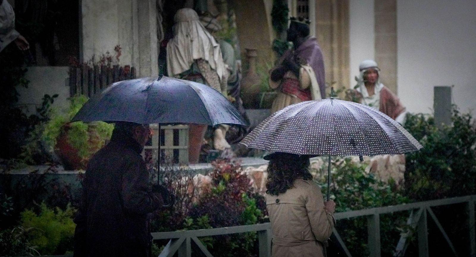 Dos personas con paragüas, paseando junto al Belén Monumental de la Alameda Cristina.