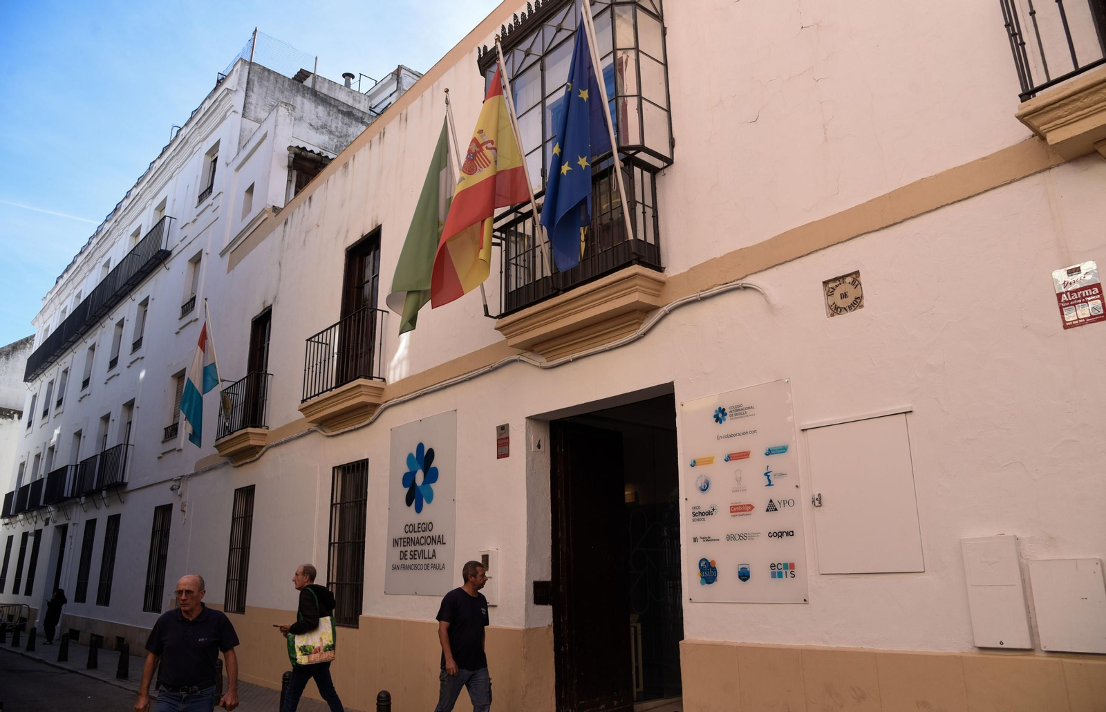 Varias personas pasan a la altura de la entrada al colegio por la calle Alcázares.