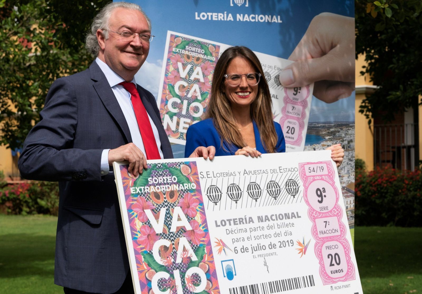 El presidente de la Sociedad Estatal de Loterías y Apuestas del Estado, junto a la alcaldesa de Ayamonte, sujetan una reproducción de un décimo de lotería del Sorteo Extraordinario de Vacaciones que se celebrará mañana en Punta del Moral.