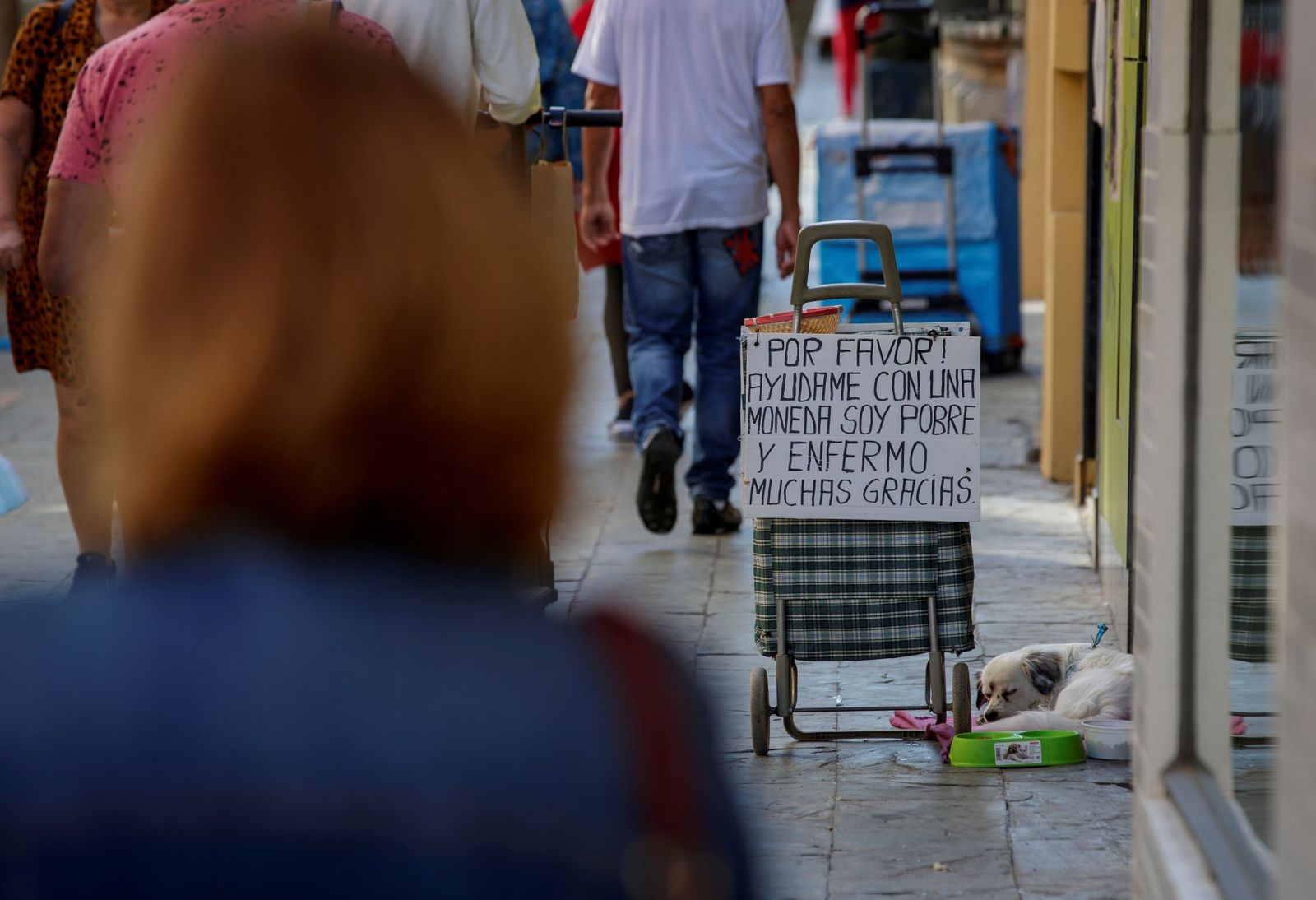 Varios viandantes pasan junto a un cartel de una persona que ejerce la mendicidad.