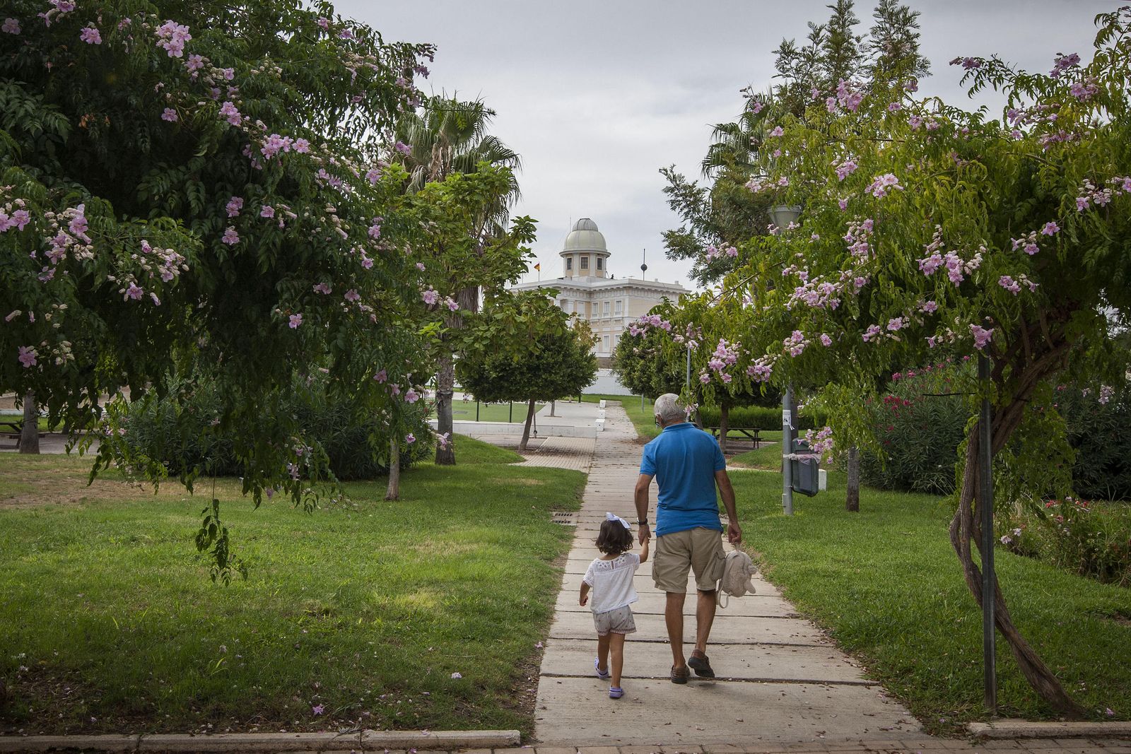 Un hombre con una pequeña de la mano pasean por los rincones del parque del Barrero, con el Observatorio al fondo.