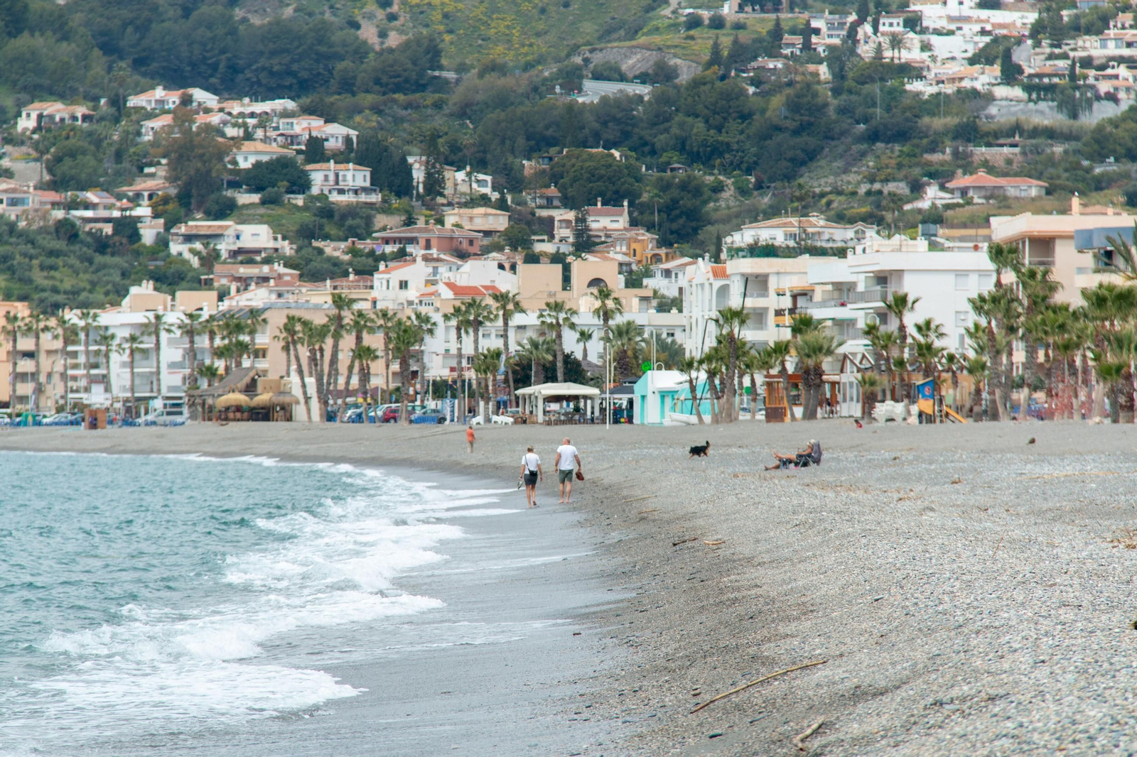 Varias personas pasean por la playa de La Herradura con las viviendas al fondo.