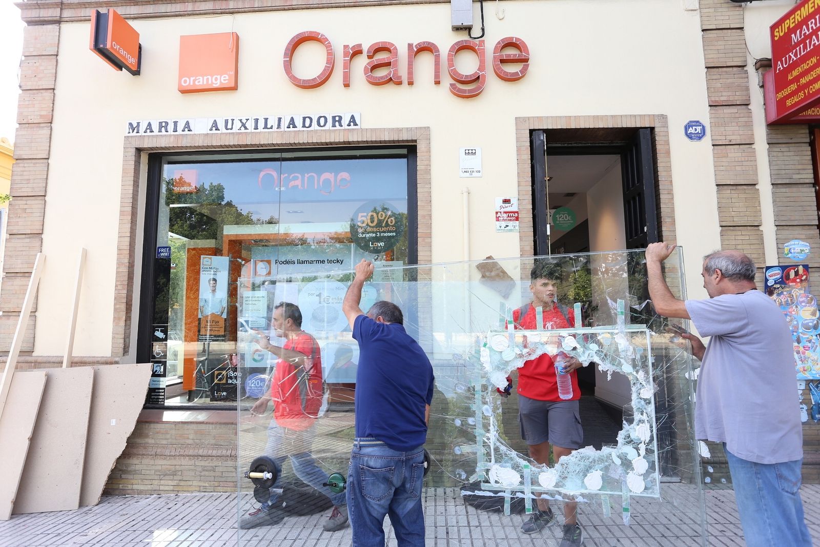 Unos operarios retiran el cristal fracturado de la tienda de Orange de María Auxiliadora.