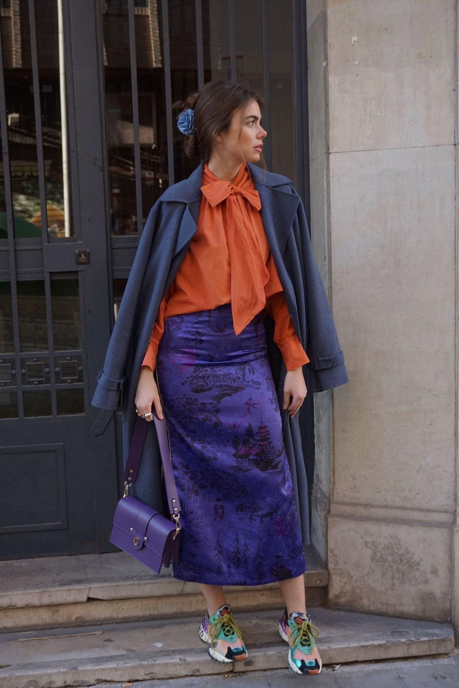 La influencer Carlota Huelva con una blusa con lazada de El armario de Paola combinada con falda midi, abrigo y deportivas.