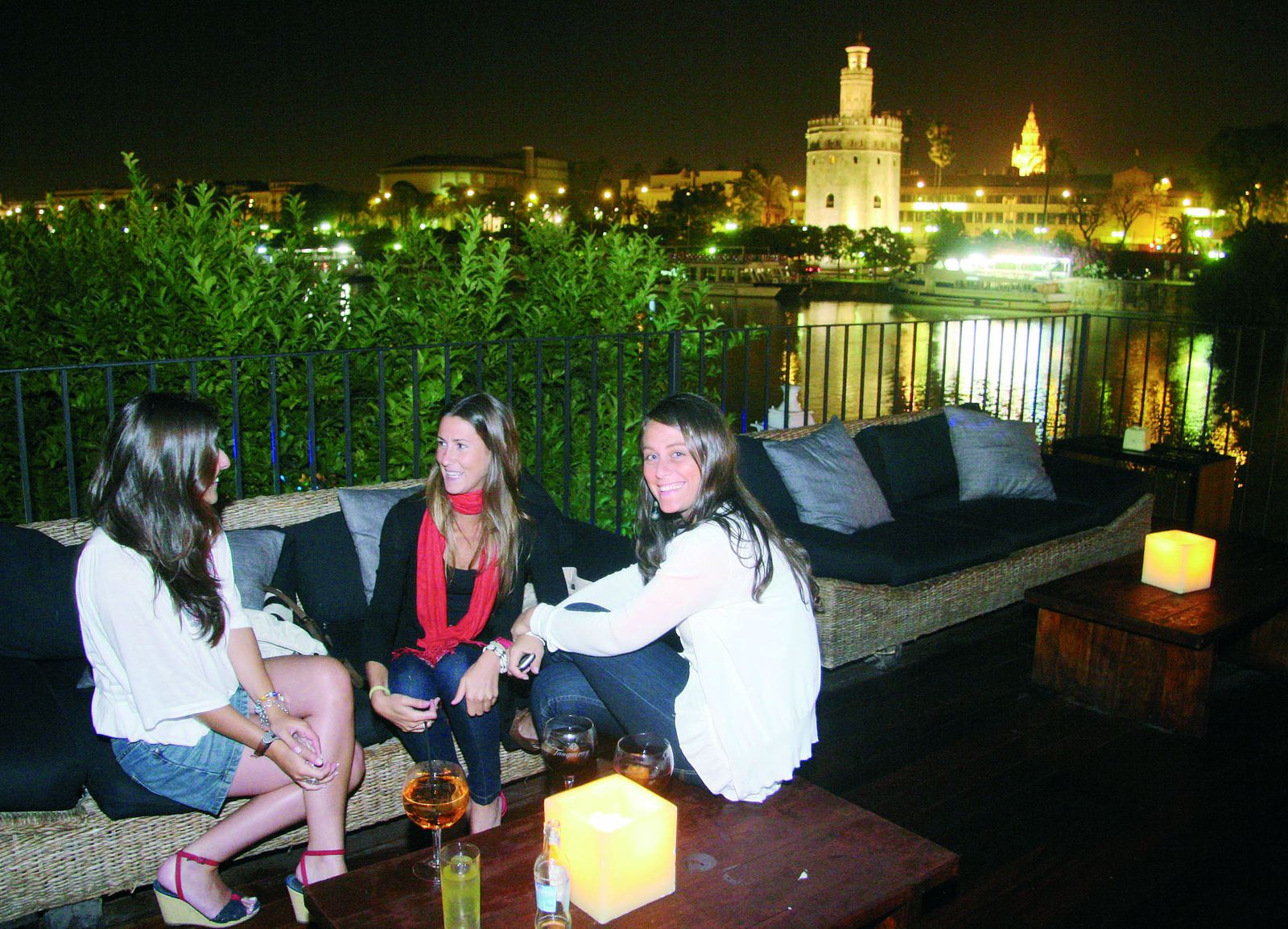 Tres jóvenes disfrutan de la noche sevillana en una terraza junto al río y con la Torre del Oro de fondo.