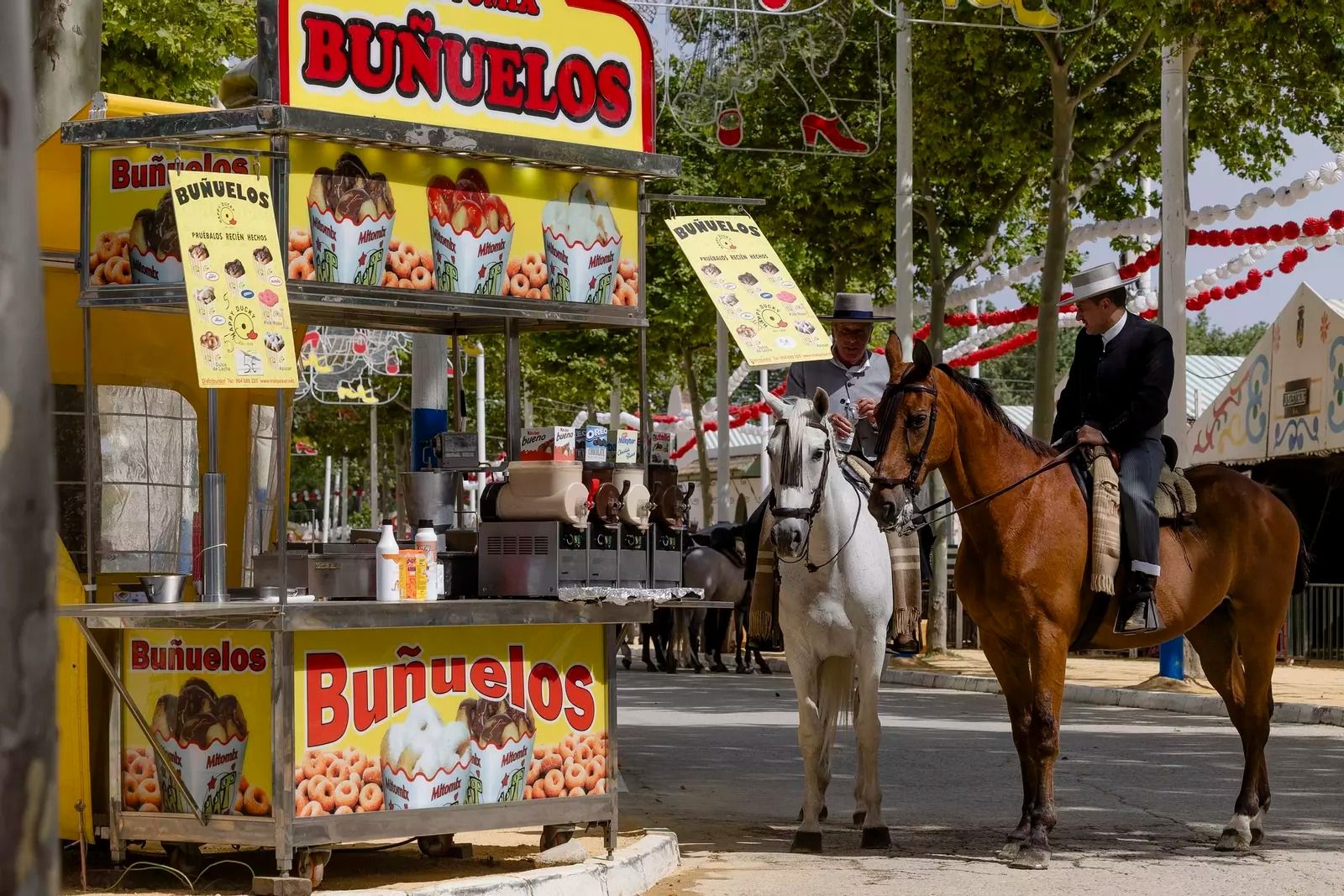 Dos jinetes a caballo en la feria de El Puerto al lado de un puesto de buñuelos