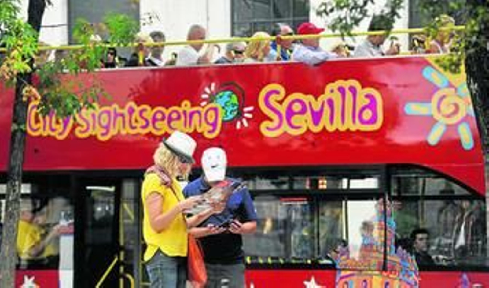 Dos foráneos consultan el mapa de la ciudad con el autobús turístico, al fondo, en la Plaza del Duque.