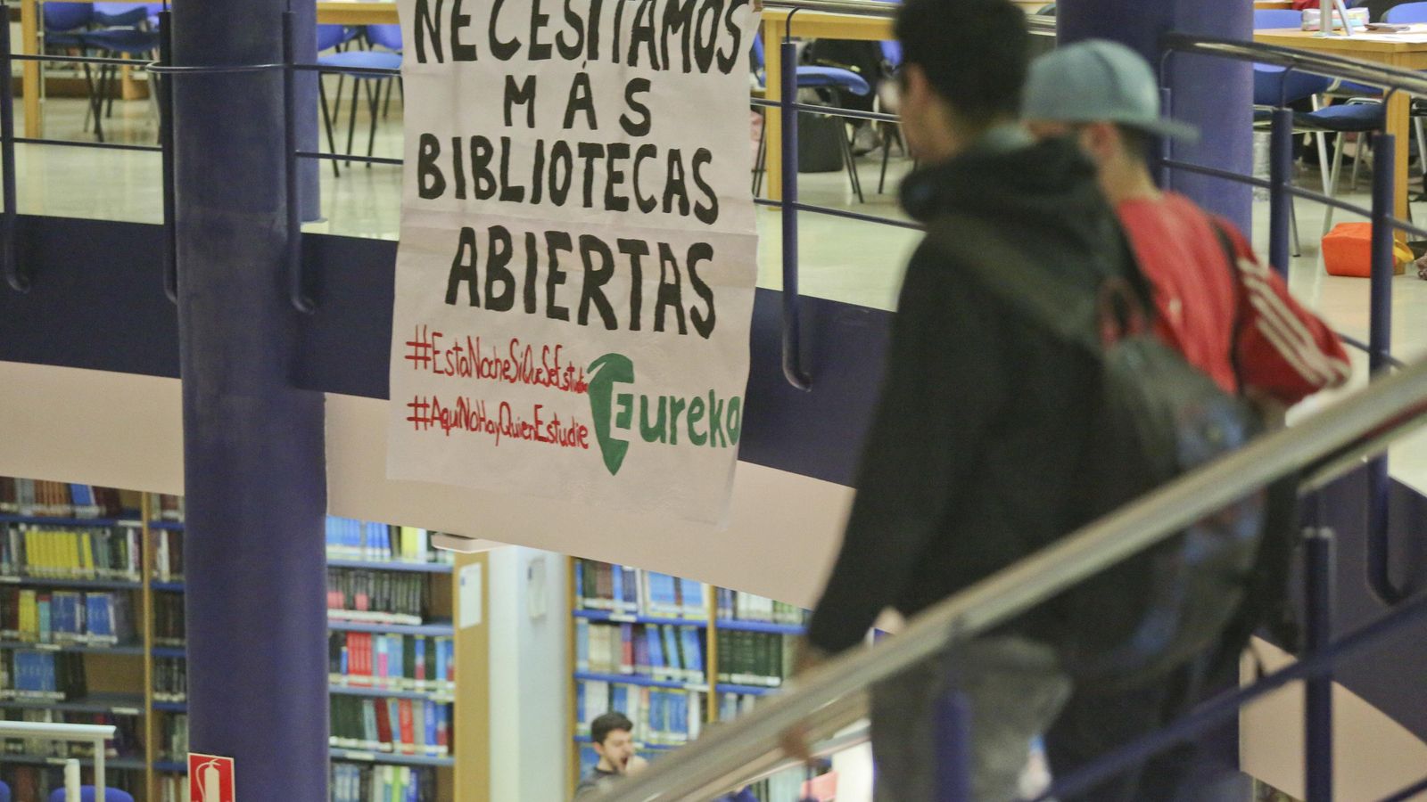 Dos estudiantes abandonan la biblioteca mientras leen la pancarta.