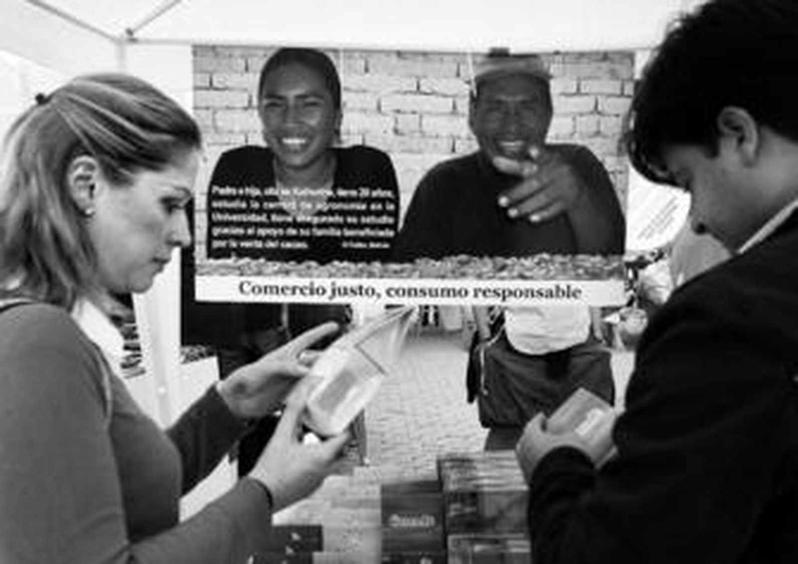 Dos visitantes de la Feria examinan algunos de los productos puestos en venta, ante un cartel de Intermón Oxfam