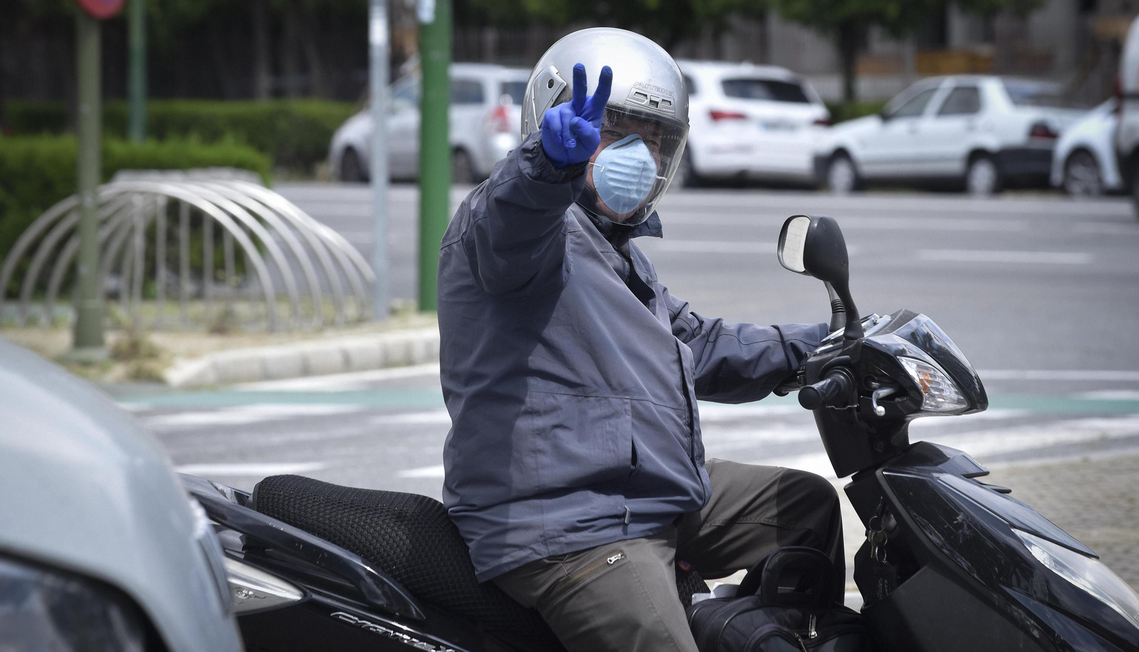 Un hombre equipado con mascarilla y guantes hace con su mano desde su moto el signo de la victoria.