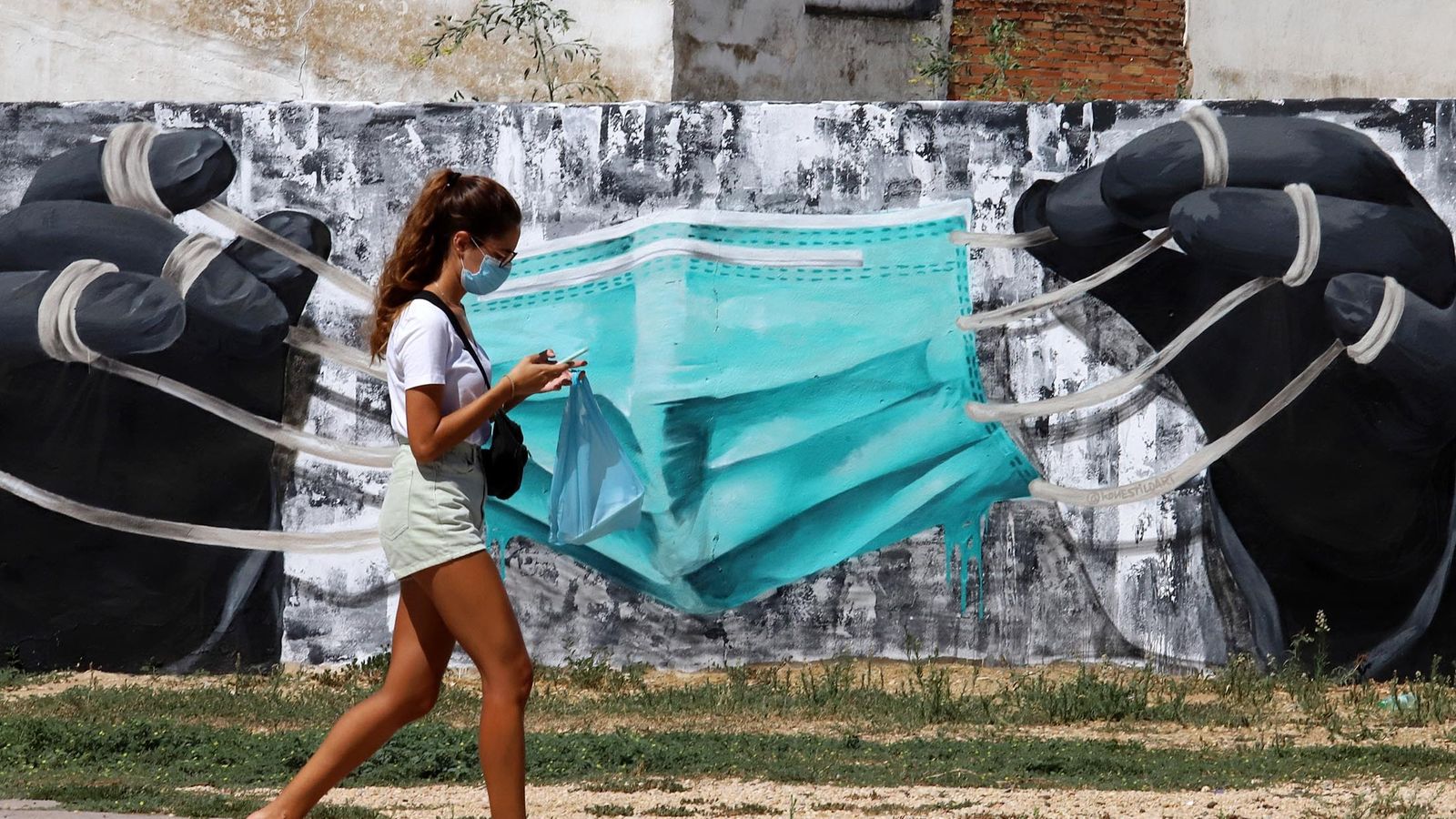 Una joven paseando por Huelva ante un muro en el que han dibujado una gran mascarilla.
