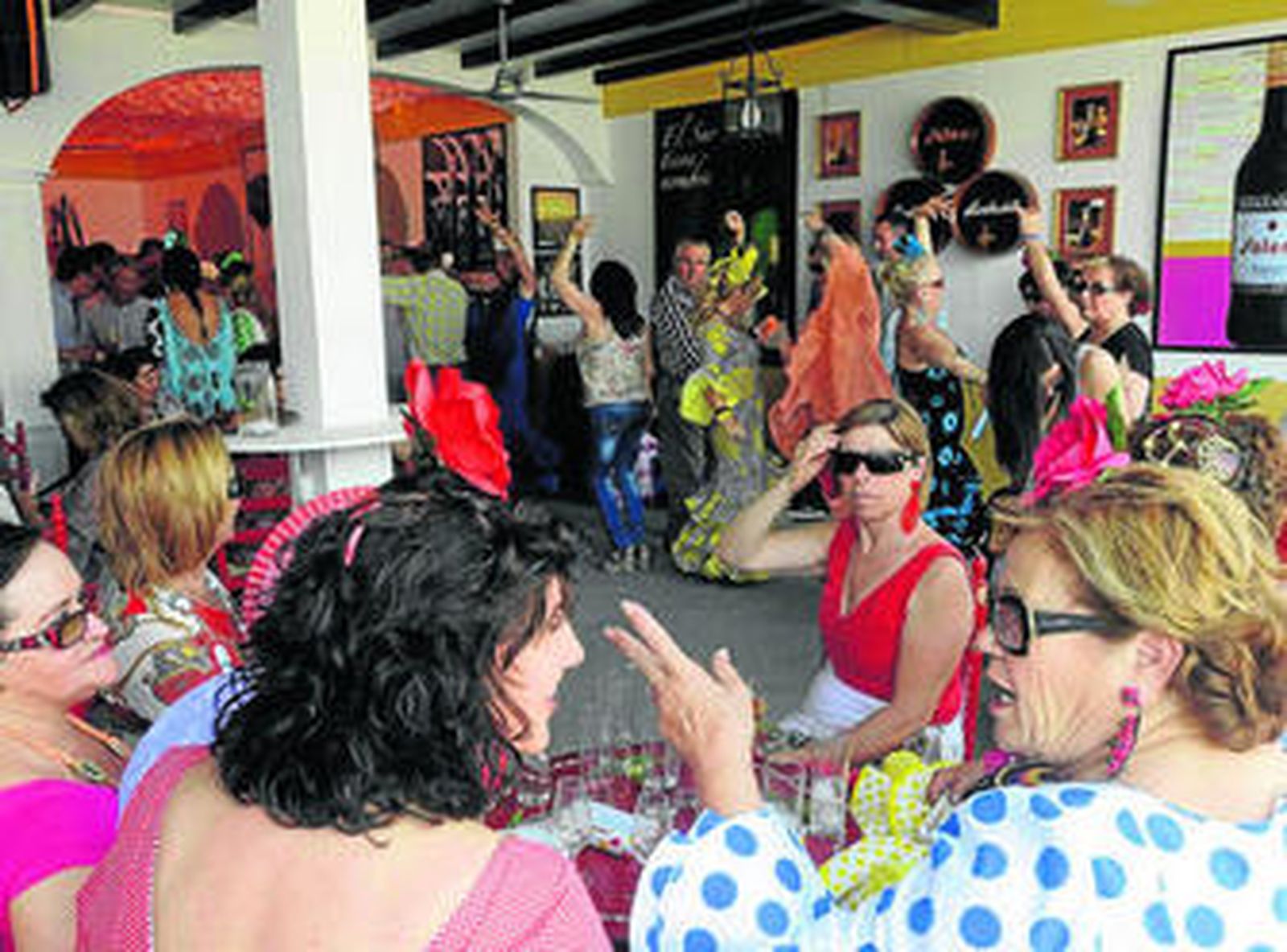Un grupo de mujeres charla mientras otras personas bailan en una concurrida caseta del recinto ferial.