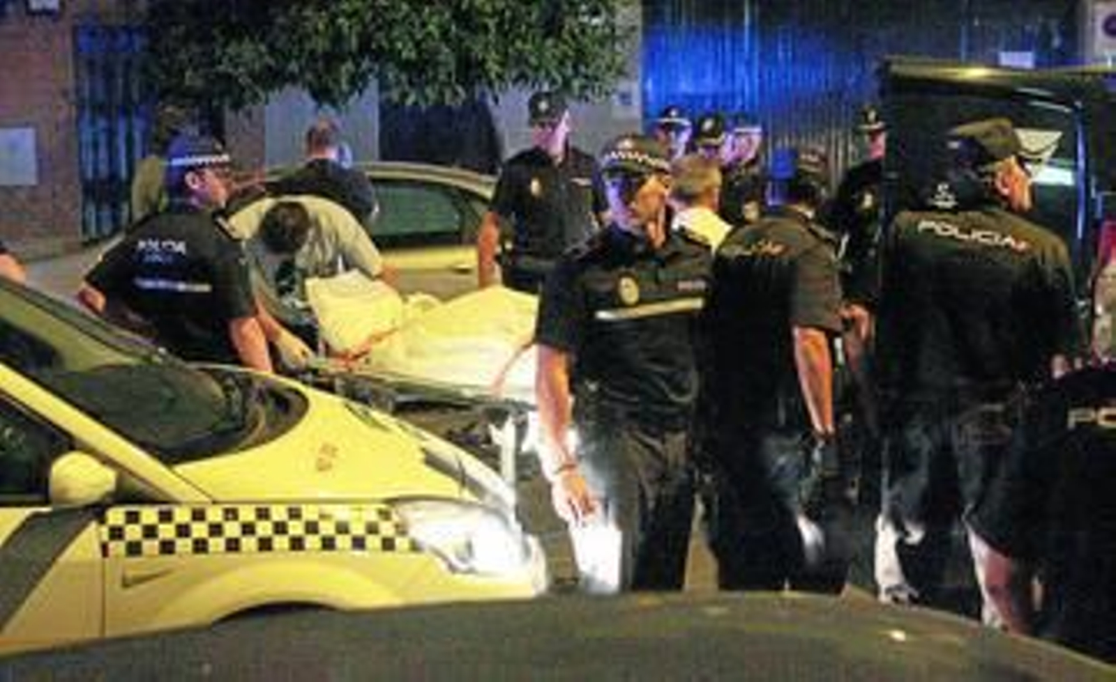 Policías locales y nacionales en la escena del crimen, mientras los operarios de los servicios funerarios trasladan el cuerpo en una camilla.