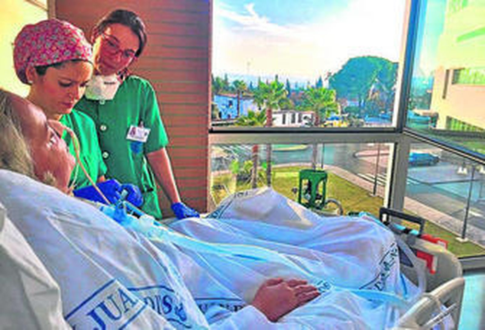 Dos enfermeras acompañan junto a un balcón a una paciente encamada.