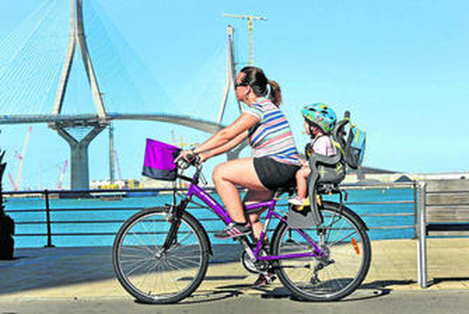 Una mujer monta en bicicleta transportando a su hijo por el Paseo de la Bahía con el segundo puente de fondo.