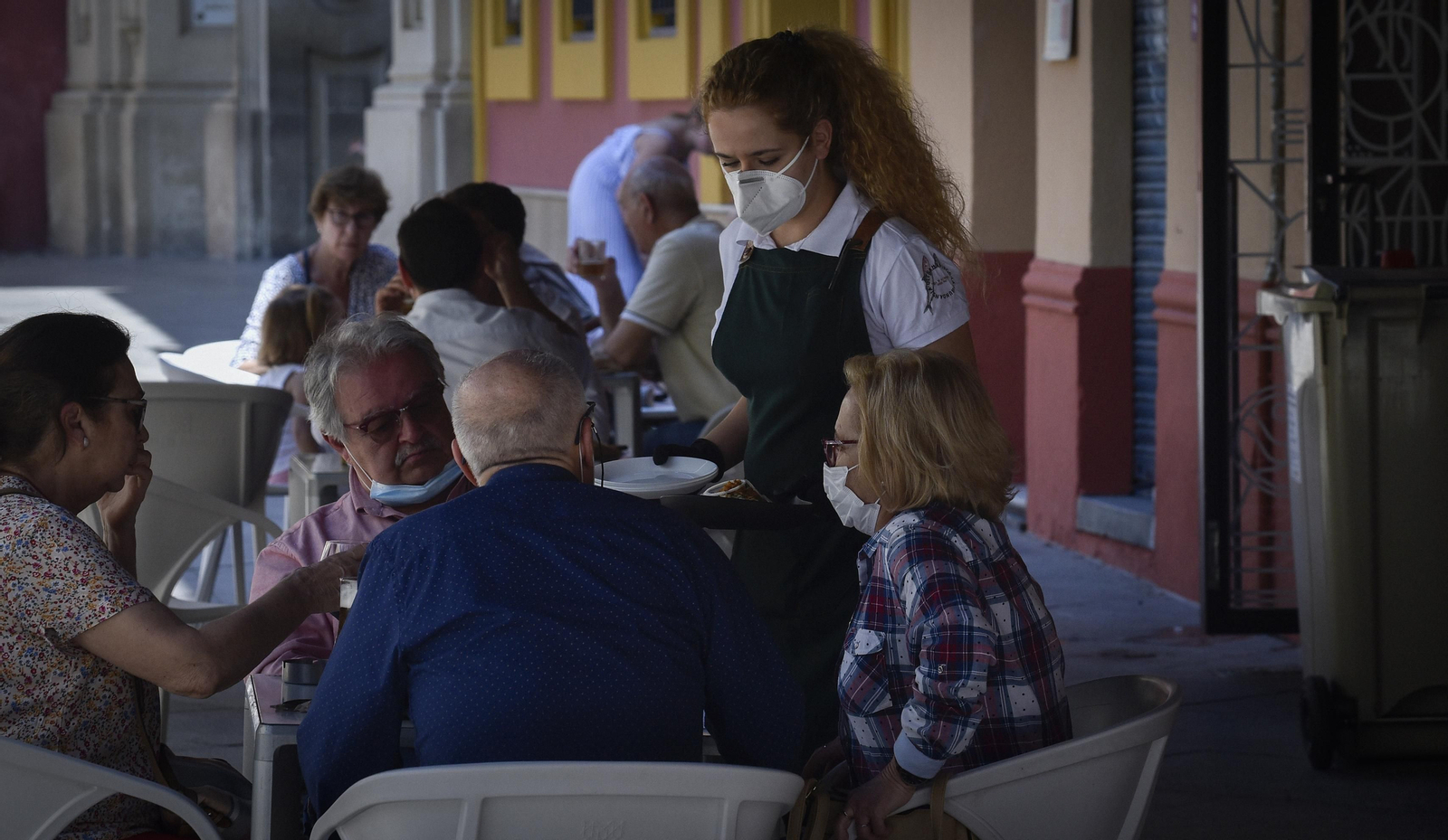 Una camarera atiende una mesa protegida por una mascarilla.
