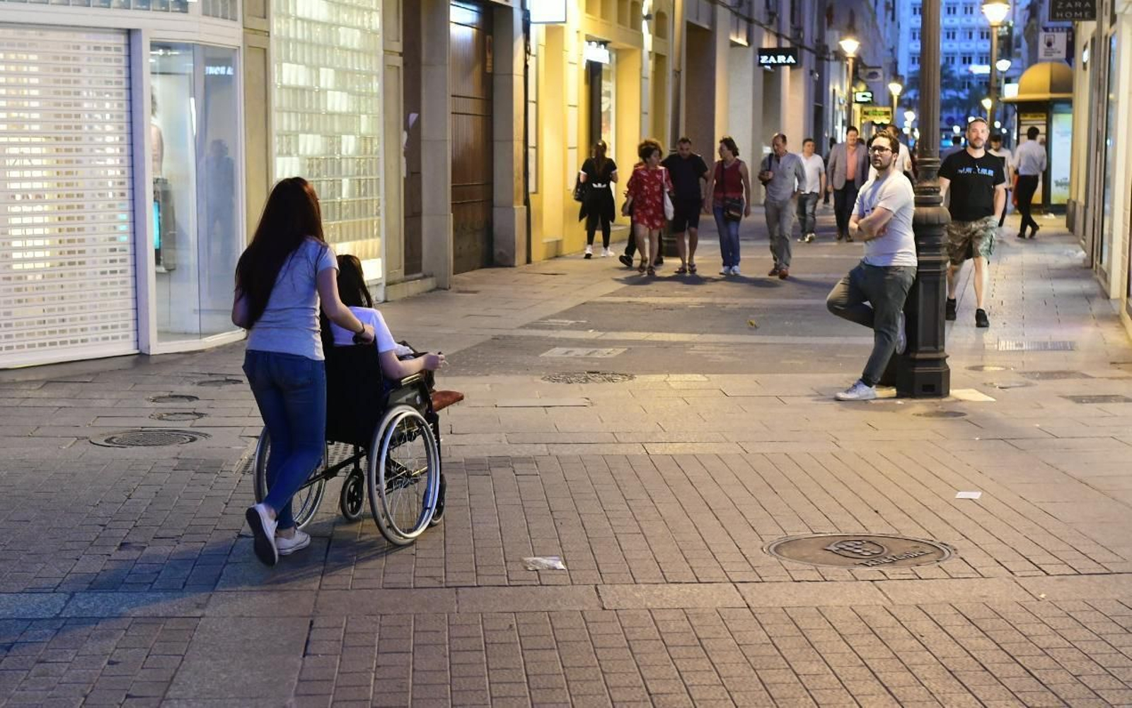 Una joven ayuda a desplazarse a otra en silla de ruedas por el centro de la ciudad.