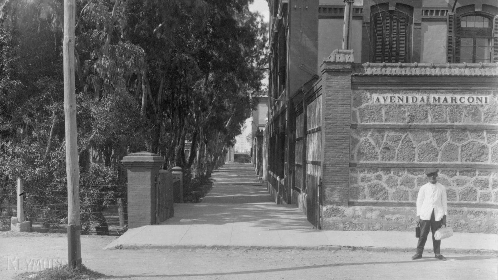 Tarjeta postal Reymundo que muestra un barbero a las puertas de los depósitos por el lado de Marconi, con las torres del Telegrafía sin Hilos al fondo.