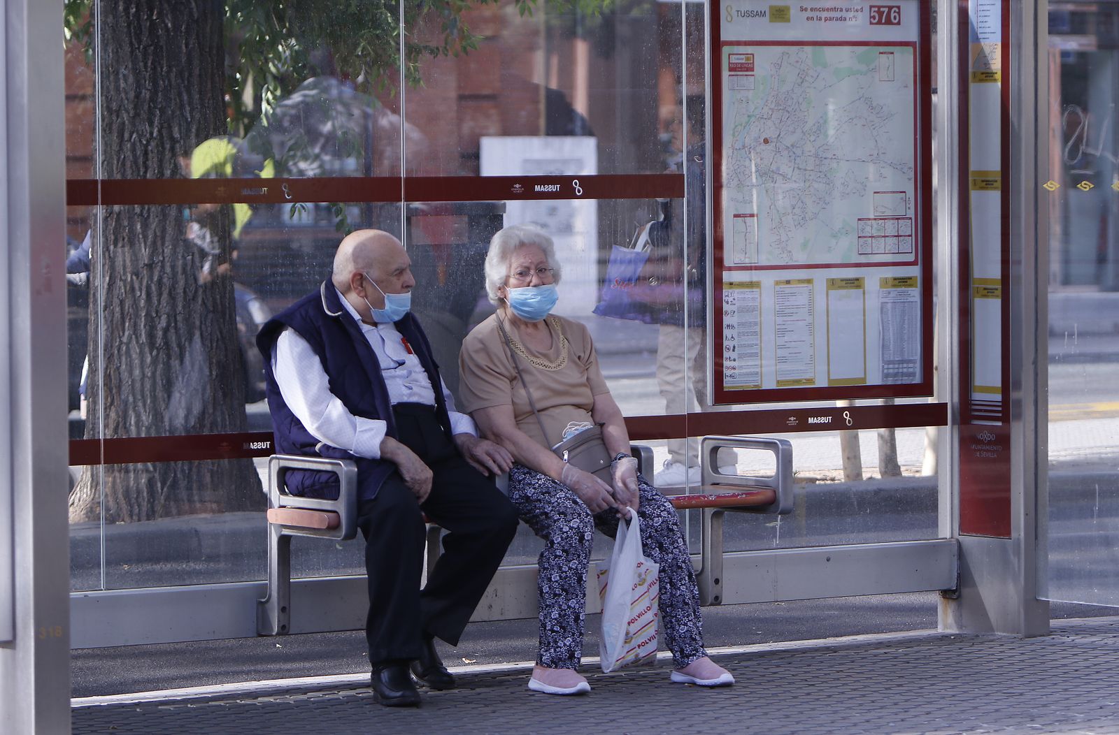Dos personas mayores con mascarilla esperan el autobús en una parada de Tussam en Sevilla.